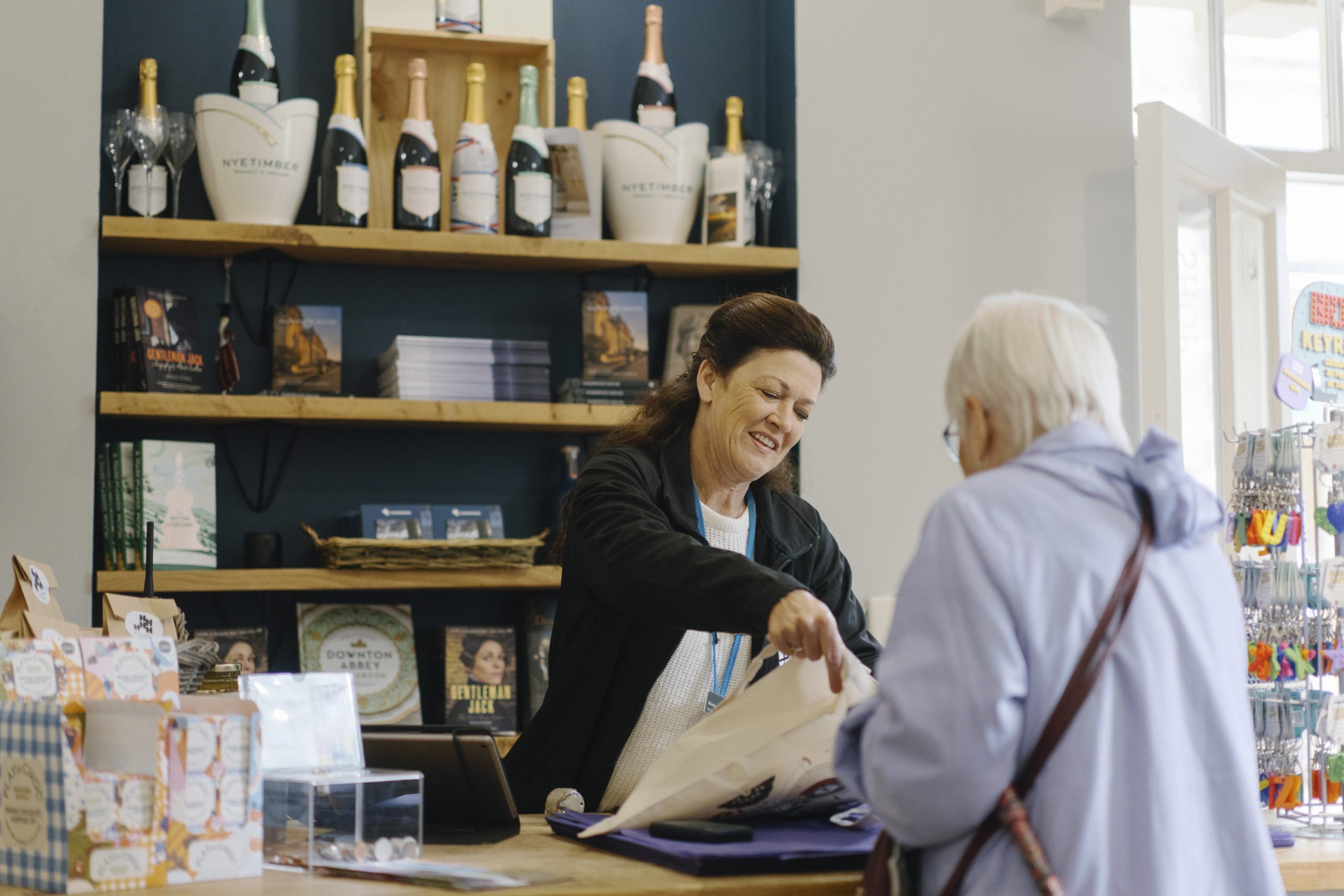 Happy Harewood Shop Assistant helping a visitor with their purchases in the Courtyard Shop. In the background, wooden shelves are filled with bottles of Nyetimber sparkling wine, books about Harewood and official merchandise from Downton Abbey, Gentleman Jack and Emmerdale - all of which was filmed at Harewood.