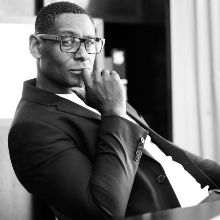 Actor David Harewood looks directly into the camera. David is wearing a well tailored suite with a crisp white shirt. He rests his elbow on a dark wooden table.