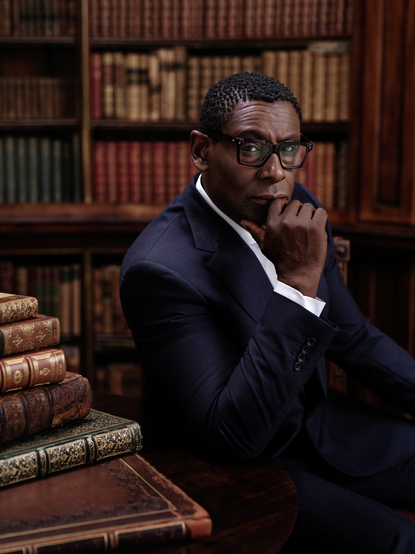 Actor David Harewood looks directly into the camera. David is wearing a well tailored navy blue suit with a crisp white shirt. He rests his elbow on a dark wooden table with old leather books piled to the side.