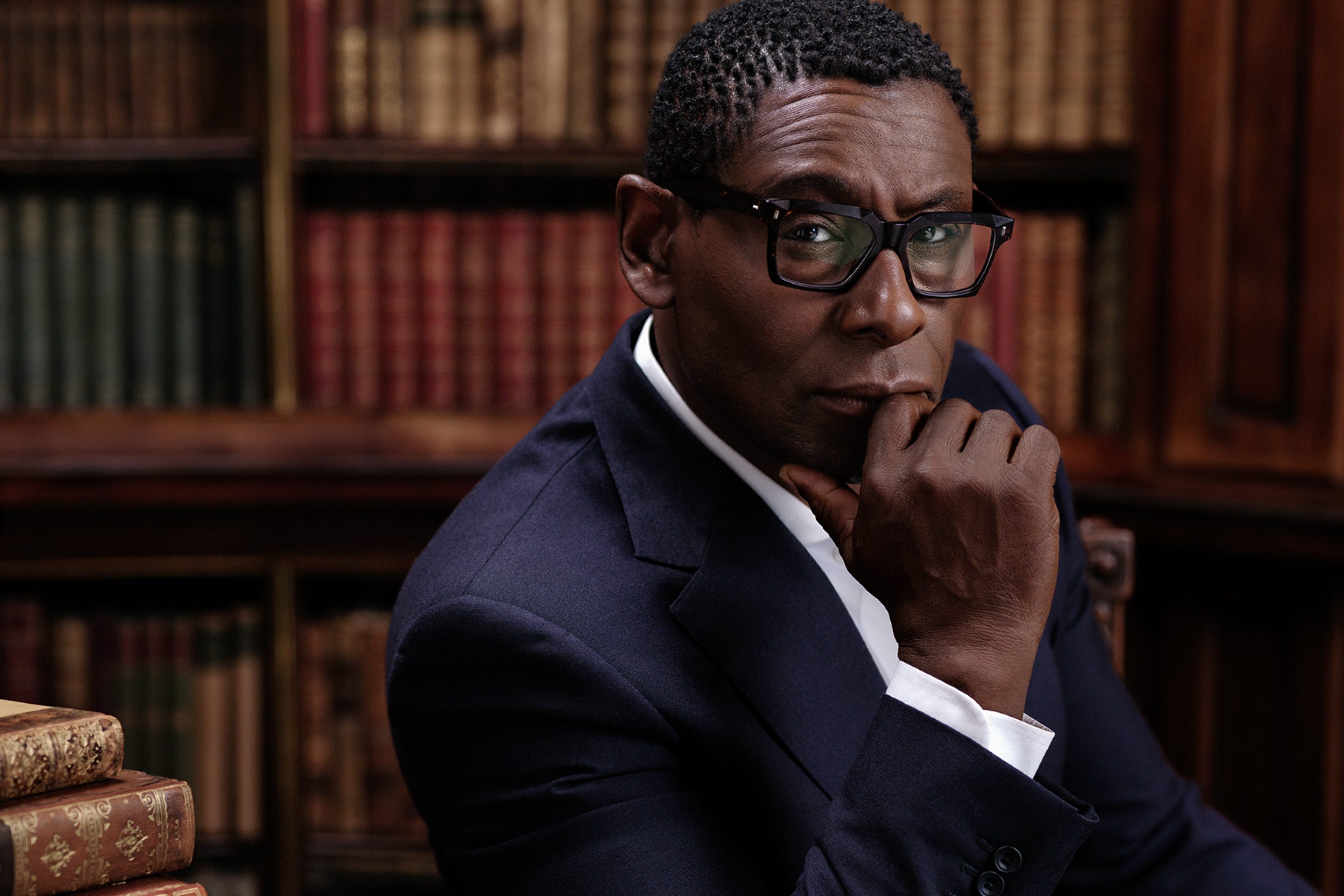 Actor David Harewood looks directly into the camera. David is wearing a well tailored navy blue suit with a crisp white shirt. He rests his elbow on a dark wooden table with old leather books piled to the side.