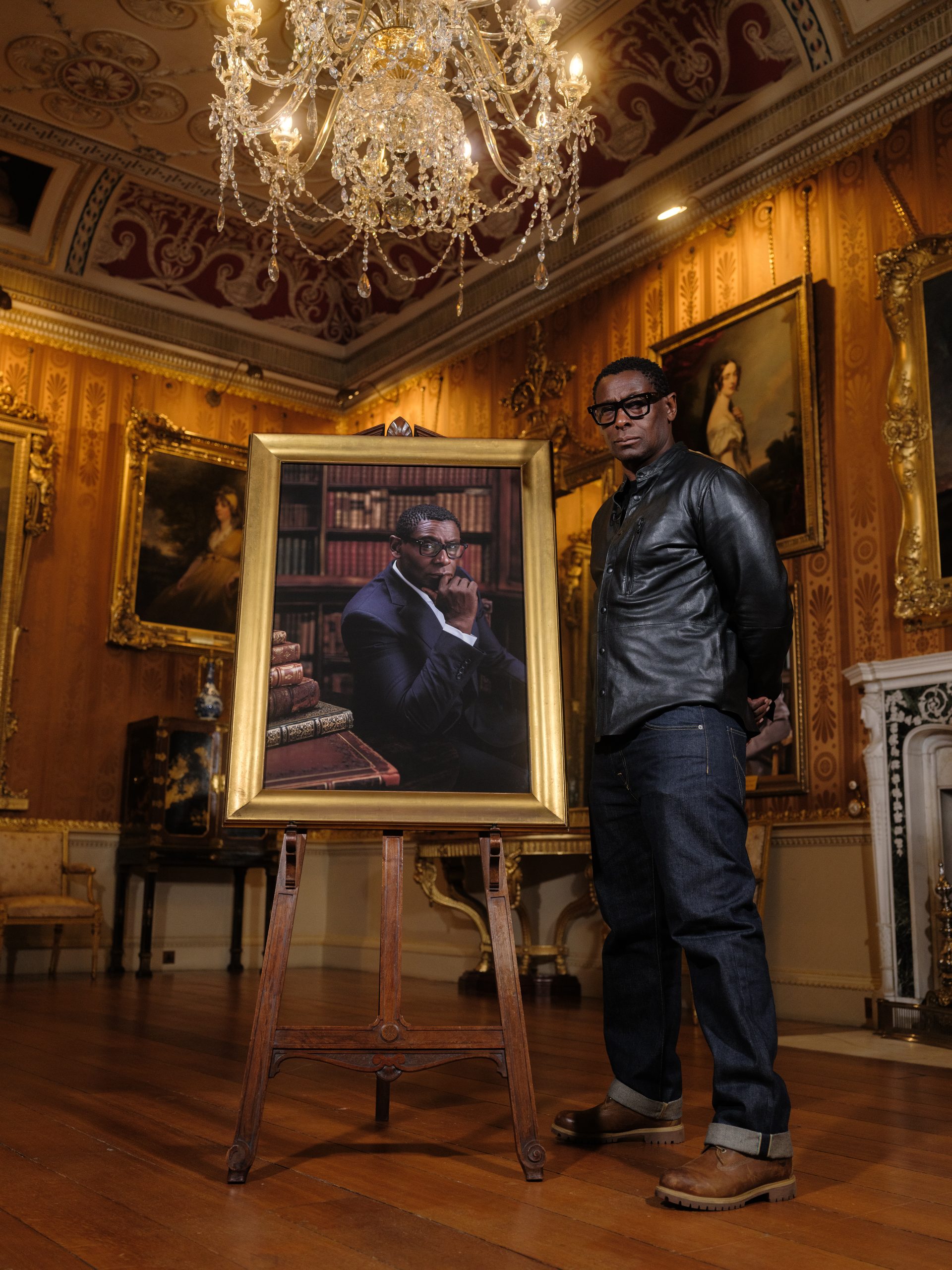 Actor David Harewood stands in the middle of the Cinnamon Drawing Room next to his portrait framed in gold on a wooden easel