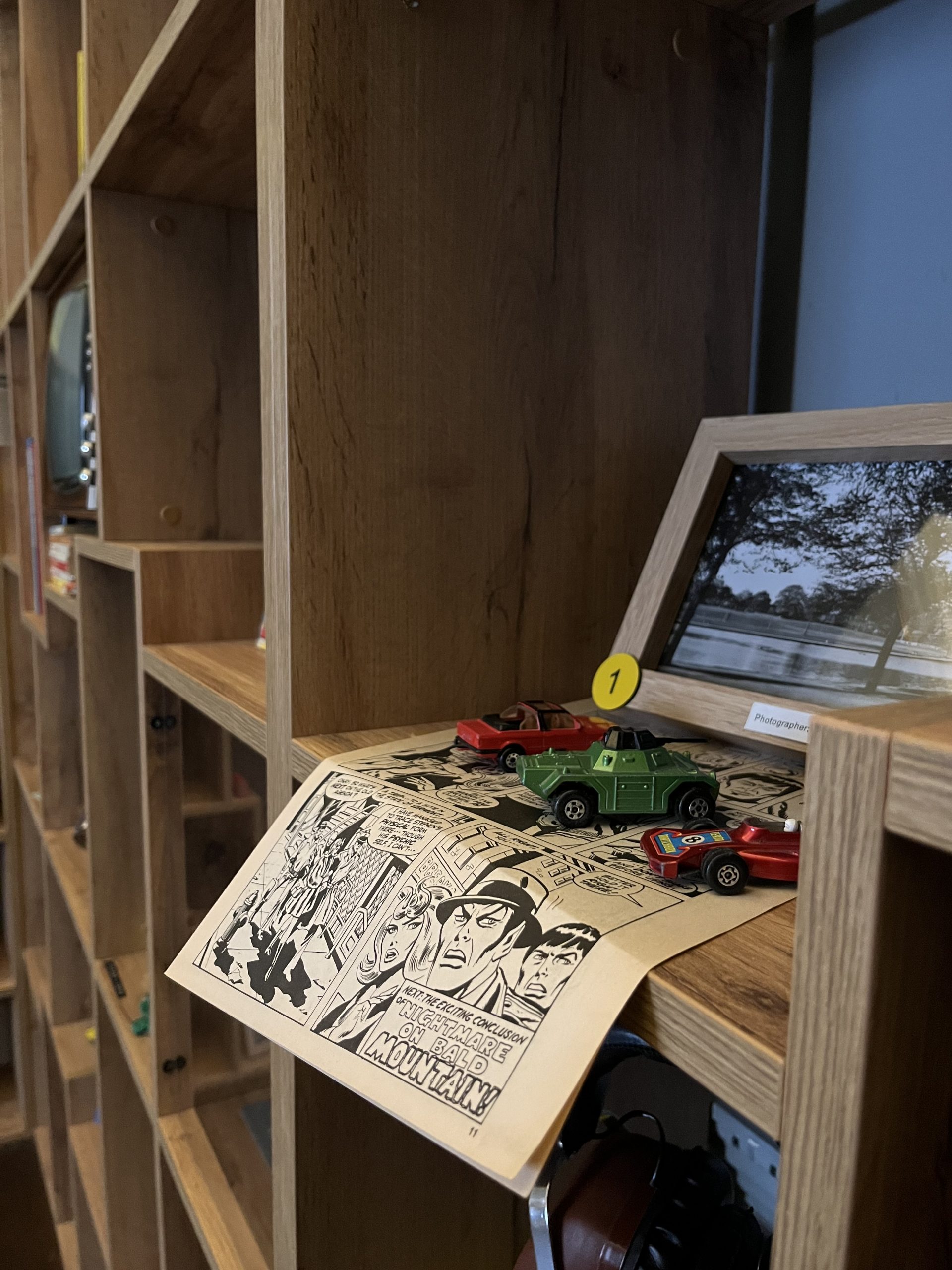 A wooden book case with black and white comic is on display with toy cars placed on top. The last scene of the comic reads "Next: the exciting conclusion of Nightmare on Bald Mountian!"