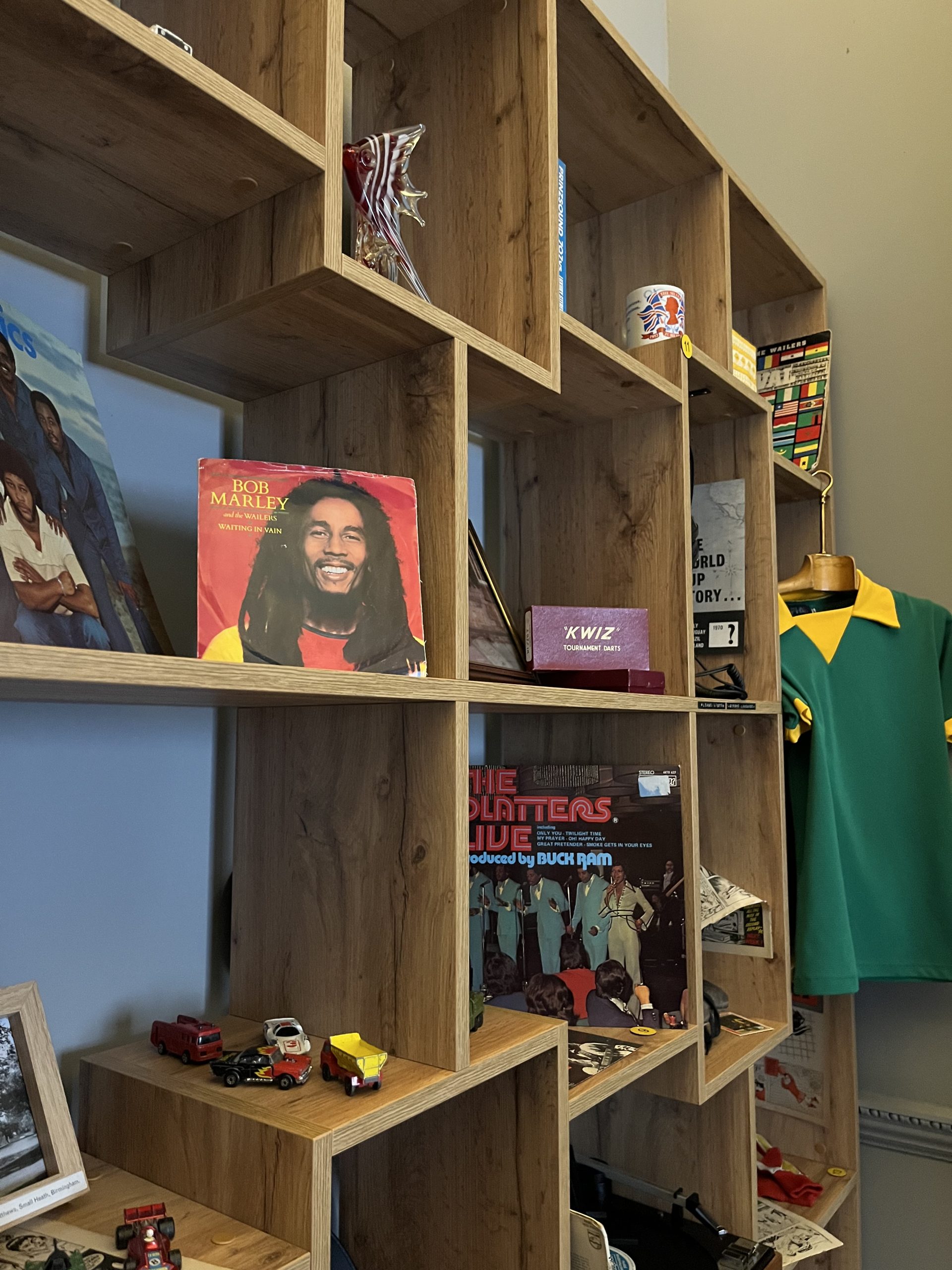 A wooden book case filled with 70s and 80s memorabilia, including Bob Marley record sleeve, toy cars, jubilee mug and comic books.