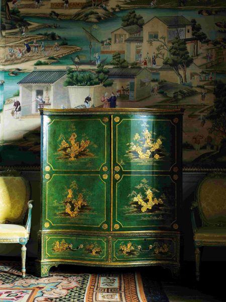 A large piece of green and gold painted furniture designed for storing clothes, displayed in front of Chinese wallpaper.