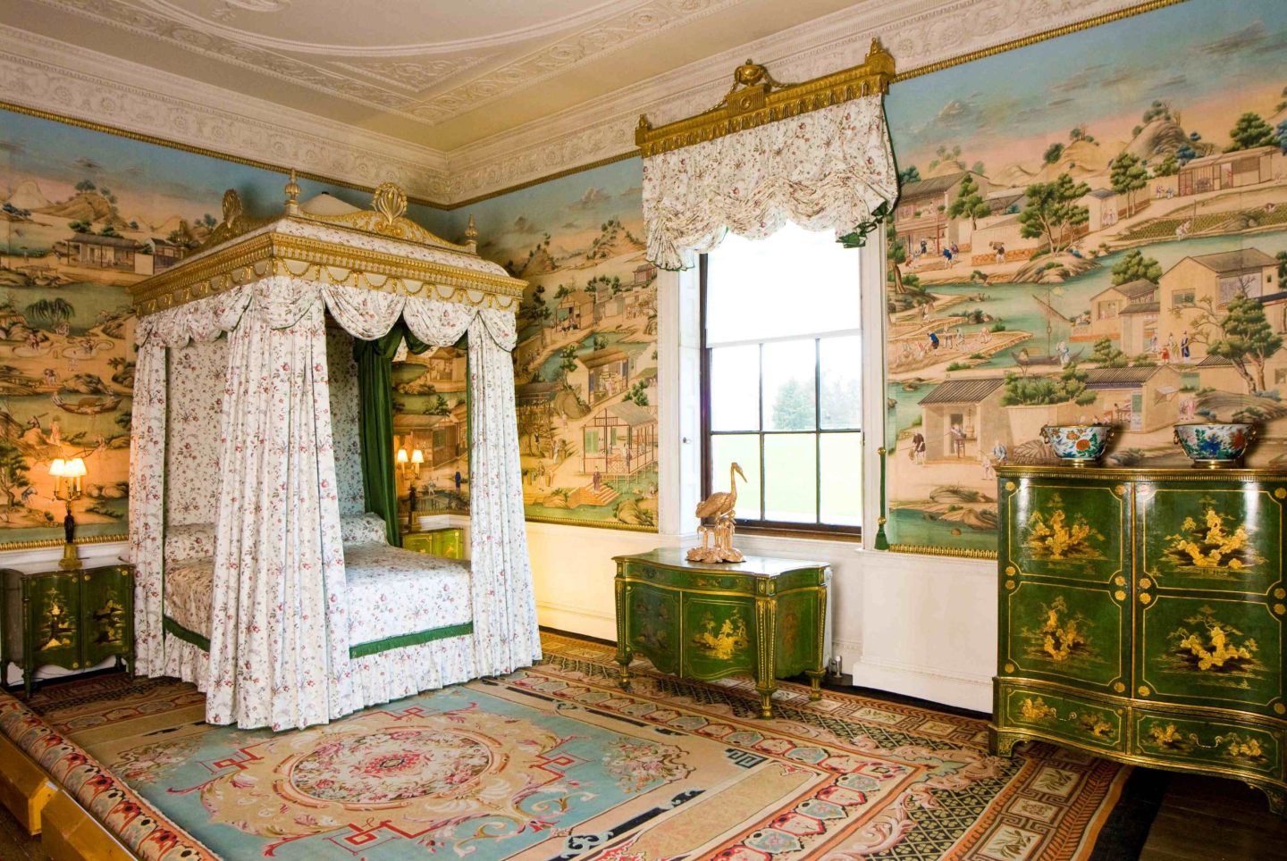 A bedroom decorated with colourful Chinese wallpaper. A large four-poster bed with white drapes is present in one corner alongside several pieces of green Chinese-style pieces of furniture.