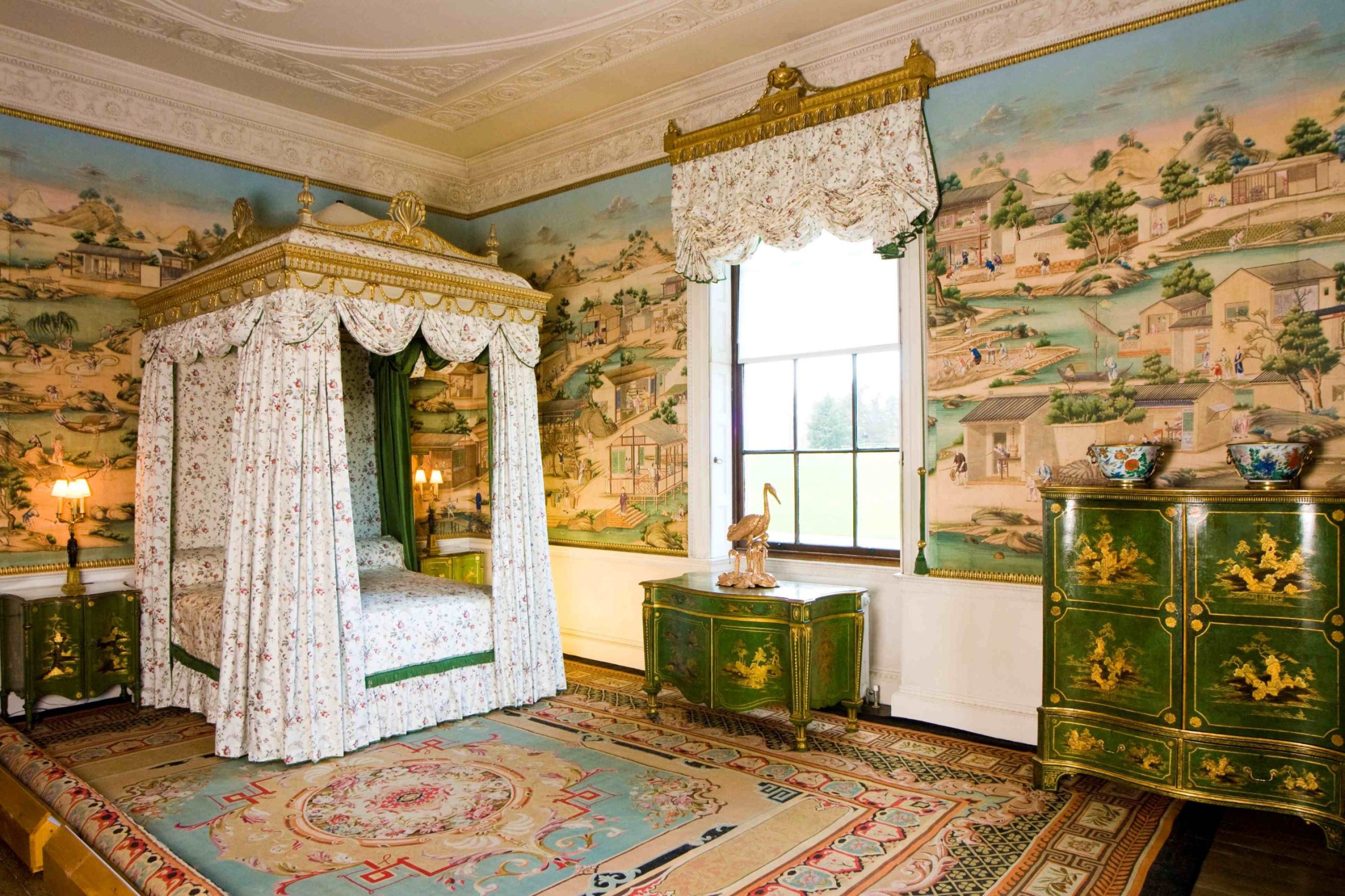 A bedroom decorated with colourful Chinese wallpaper. A large four-poster bed with white drapes is present in one corner alongside several pieces of green Chinese-style pieces of furniture.