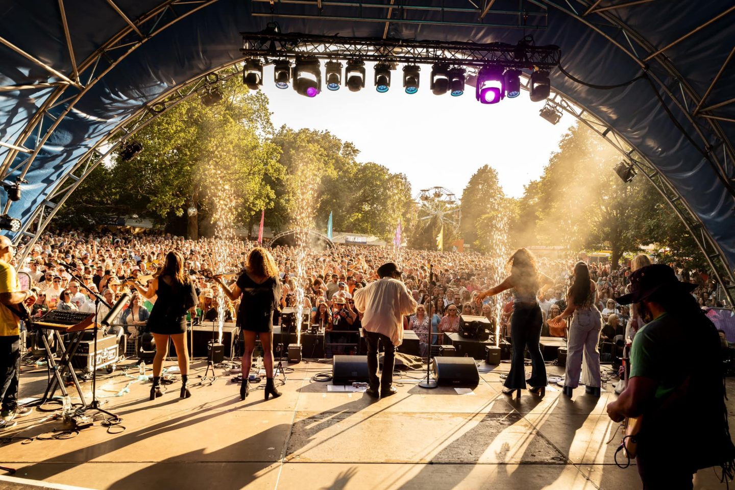 5 band members perform on stage in front of a huge crowd of people. Fireworks shoot up from the front of the stage as the crowd cheer happily.