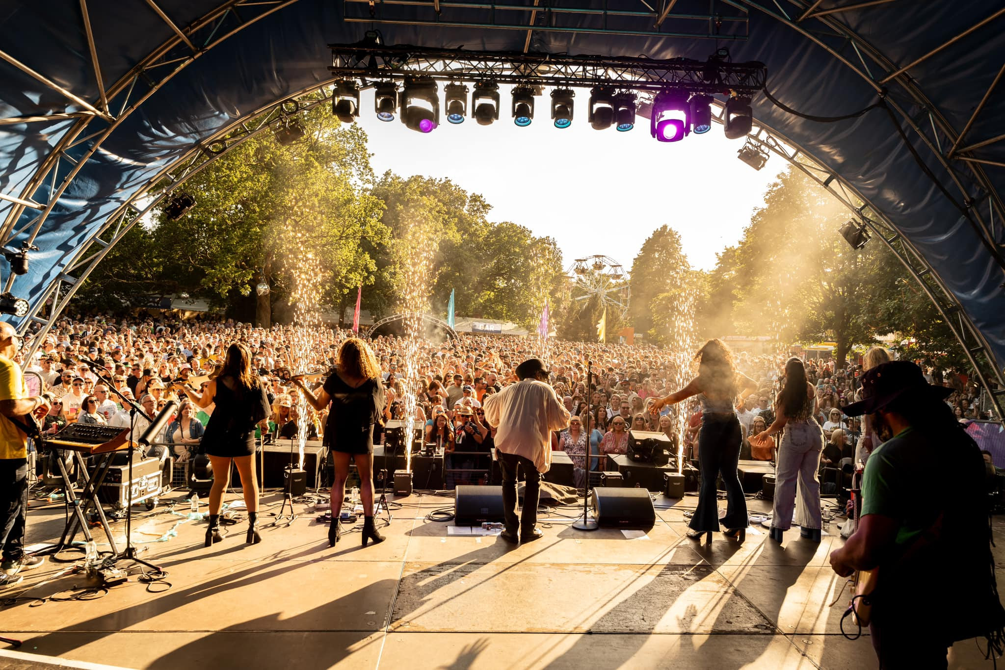 5 band members perform on stage in front of a huge crowd of people. Fireworks shoot up from the front of the stage as the crowd cheer happily.