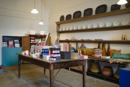 Shop interior with a rustic wooden table displaying colourful books and stationery. Shelves hold plates, woven baskets, and vibrant textiles.