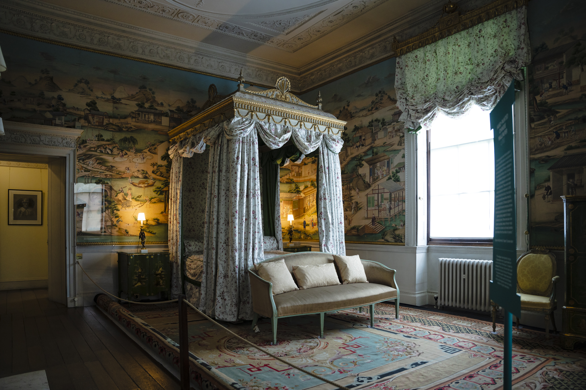 A large gilded four poster bed designed by Thomas Chippendale stands in the centre of a decorative rug with a sage and gold sofa placed at the foot of the bed. The walls are decorated with hand painted chinese wallpaper and the soft glow of bed side lamps gently light the room.