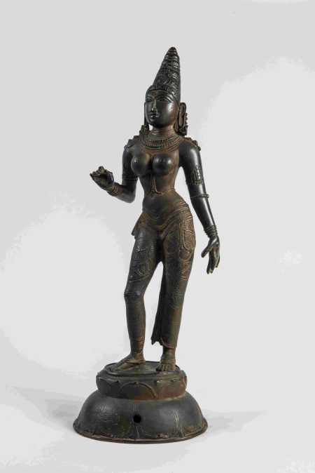 A full-length bronze sculpture of the Hindu goddess Parvati. She wears a conical head-dress and jewellery. One knee and arm is bent.