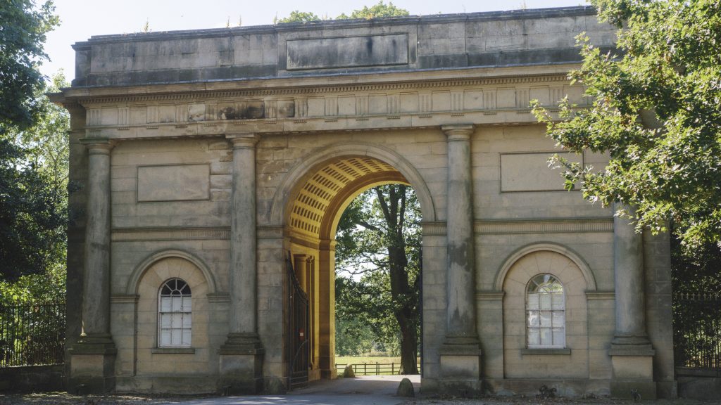 An elegant stone arched gateway at Harewood House, featuring classical Palladian design with arched openings, tall columns, and decorative detailing. The central arch frames a view of trees and a sunlit path, while smaller arched windows flank either side. The structure is surrounded by greenery, adding to its timeless charm.