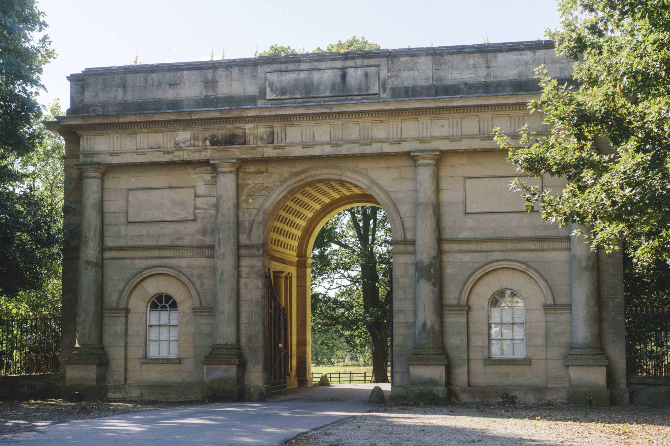 An elegant stone arched gateway at Harewood House, featuring classical Palladian design with arched openings, tall columns, and decorative detailing. The central arch frames a view of trees and a sunlit path, while smaller arched windows flank either side. The structure is surrounded by greenery, adding to its timeless charm.