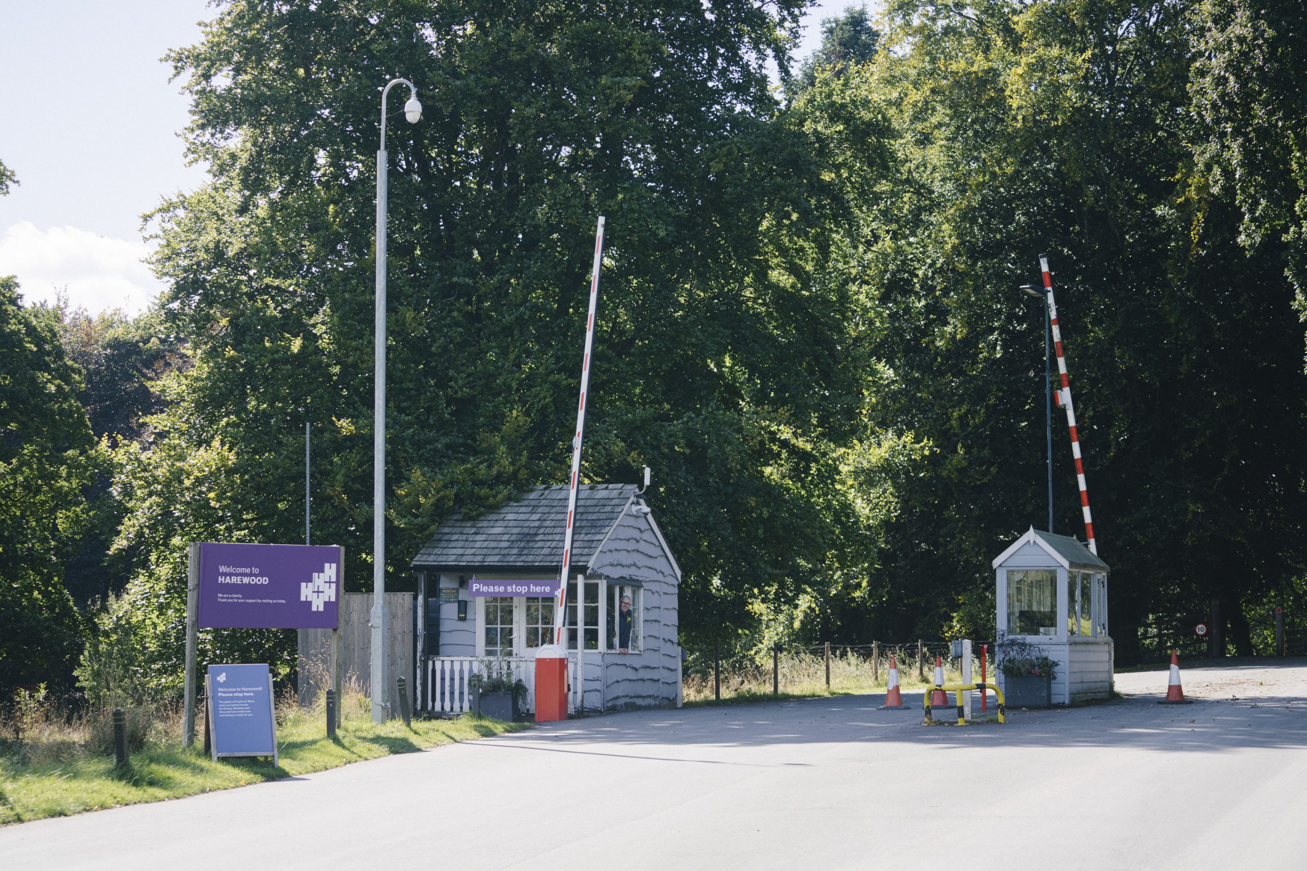 The entrance to Harewood House, featuring two small gatehouses with white wooden exteriors and red-and-white barrier arms. A purple welcome sign on the left displays the Harewood logo, set against a backdrop of tall, leafy trees and a bright blue sky. Traffic cones and flower planters add detail to the scene.