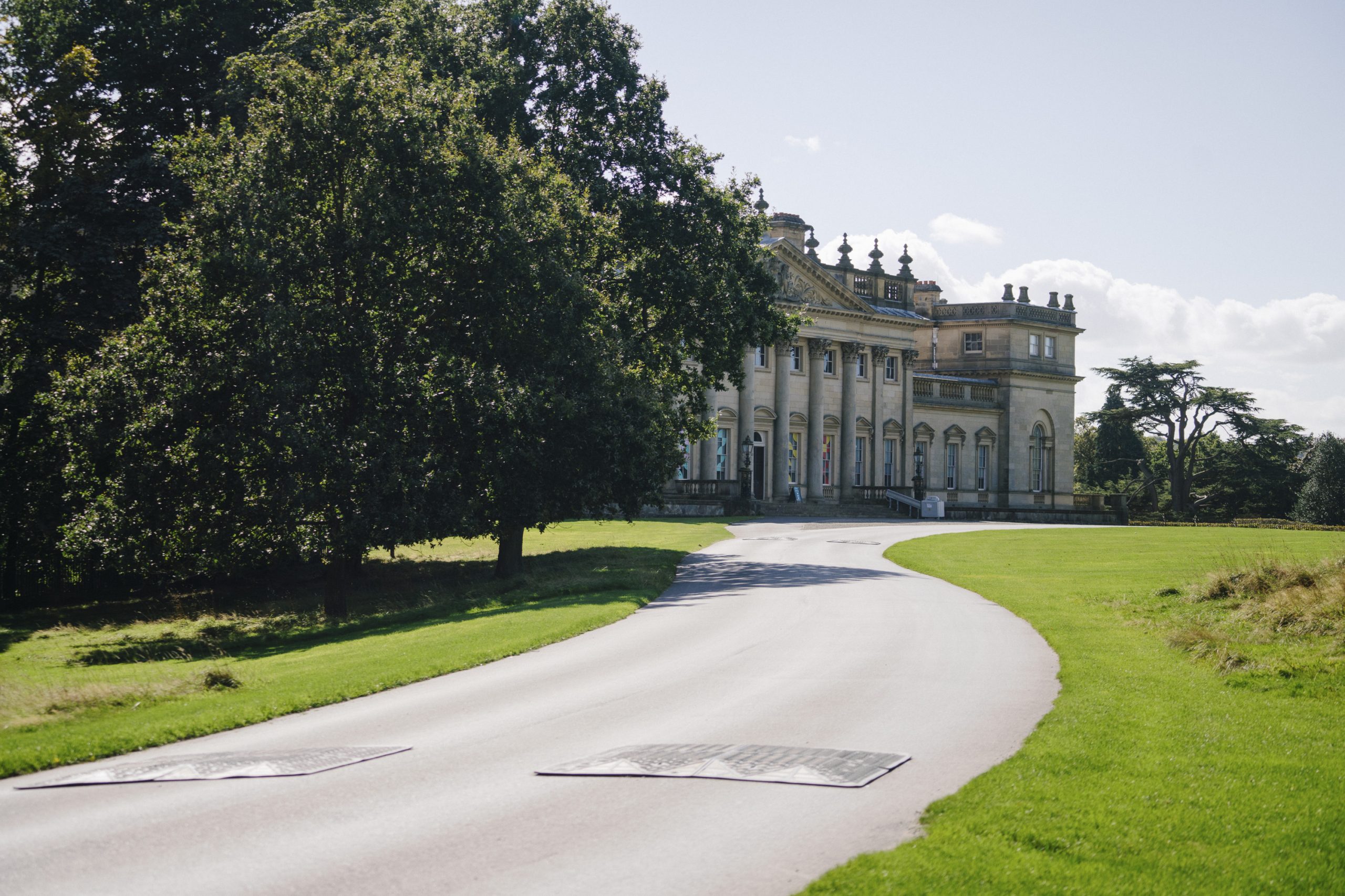 A long paved drives winds round past the front of Harewood House on the left hand side. Trees line the grass on one side, with open lawn on the the other.