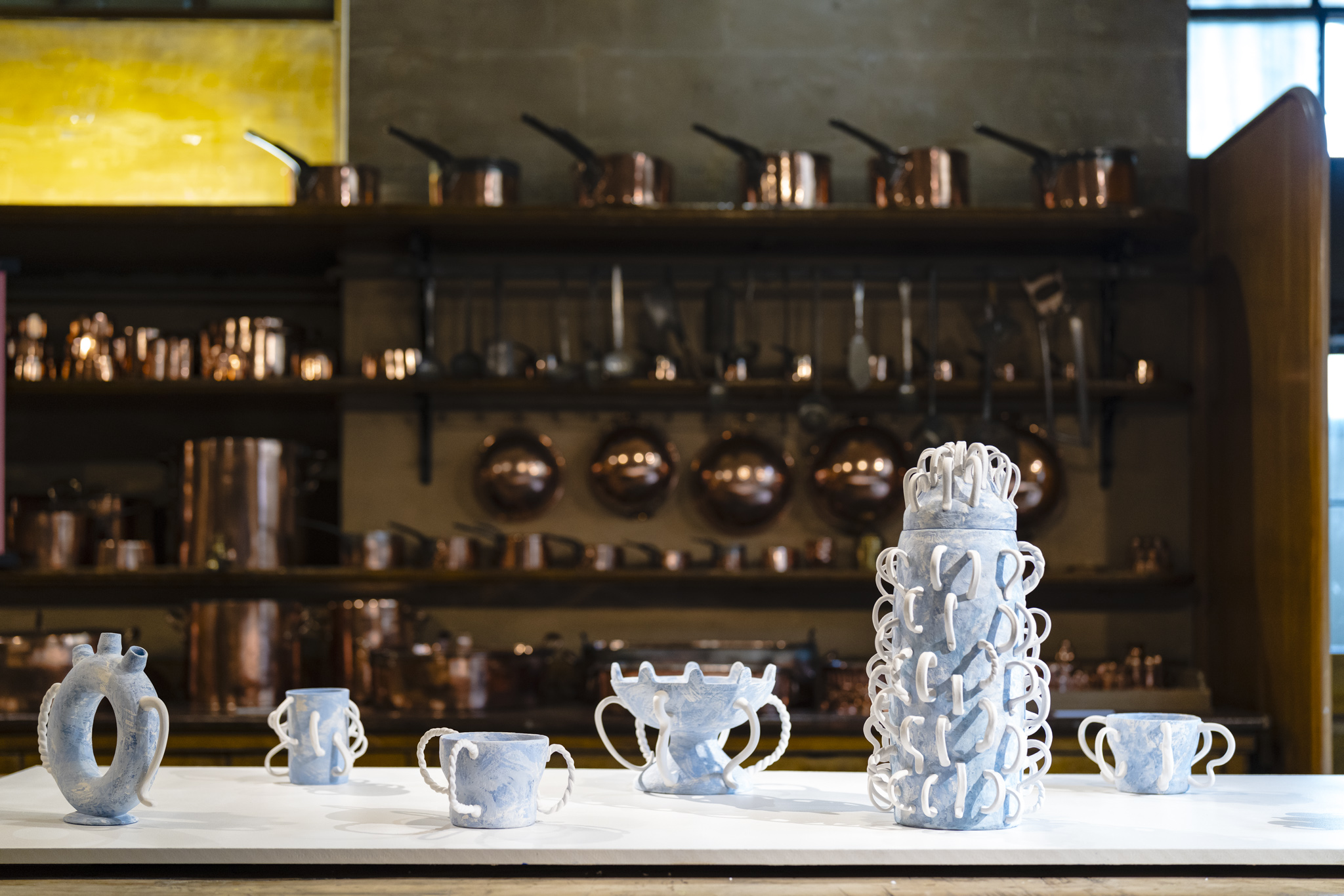 A collection of sculptural drinking vessels and tableware displayed on a countertop in the Old Kitchen. The pieces are crafted with a light blue textured finish and adorned with twisted white handles, showcasing a mix of shapes and designs. In the foreground, a tall cylindrical vessel with multiple handles stands out, surrounded by smaller cups and bowls with unique, artistic forms. In the background, copper pots, pans, and utensils hang on shelves and racks, creating a warm, traditional atmosphere.