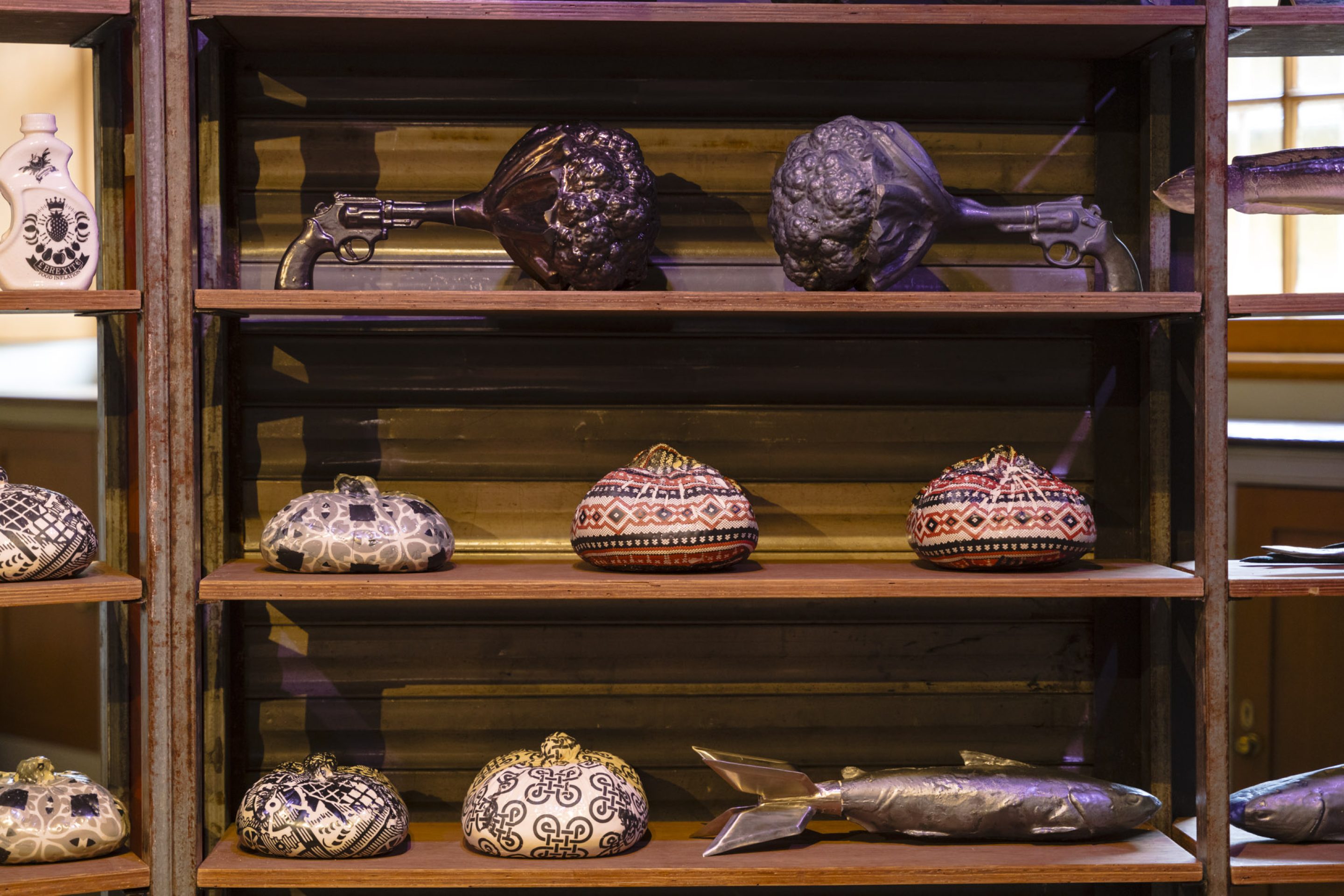 Three wooden shelves displaying ceramic objects. On the top shelf, a figure of a gun shooting cauliflowers instead of bullets. On the bottom shelves, a fish in the shape of a missile.