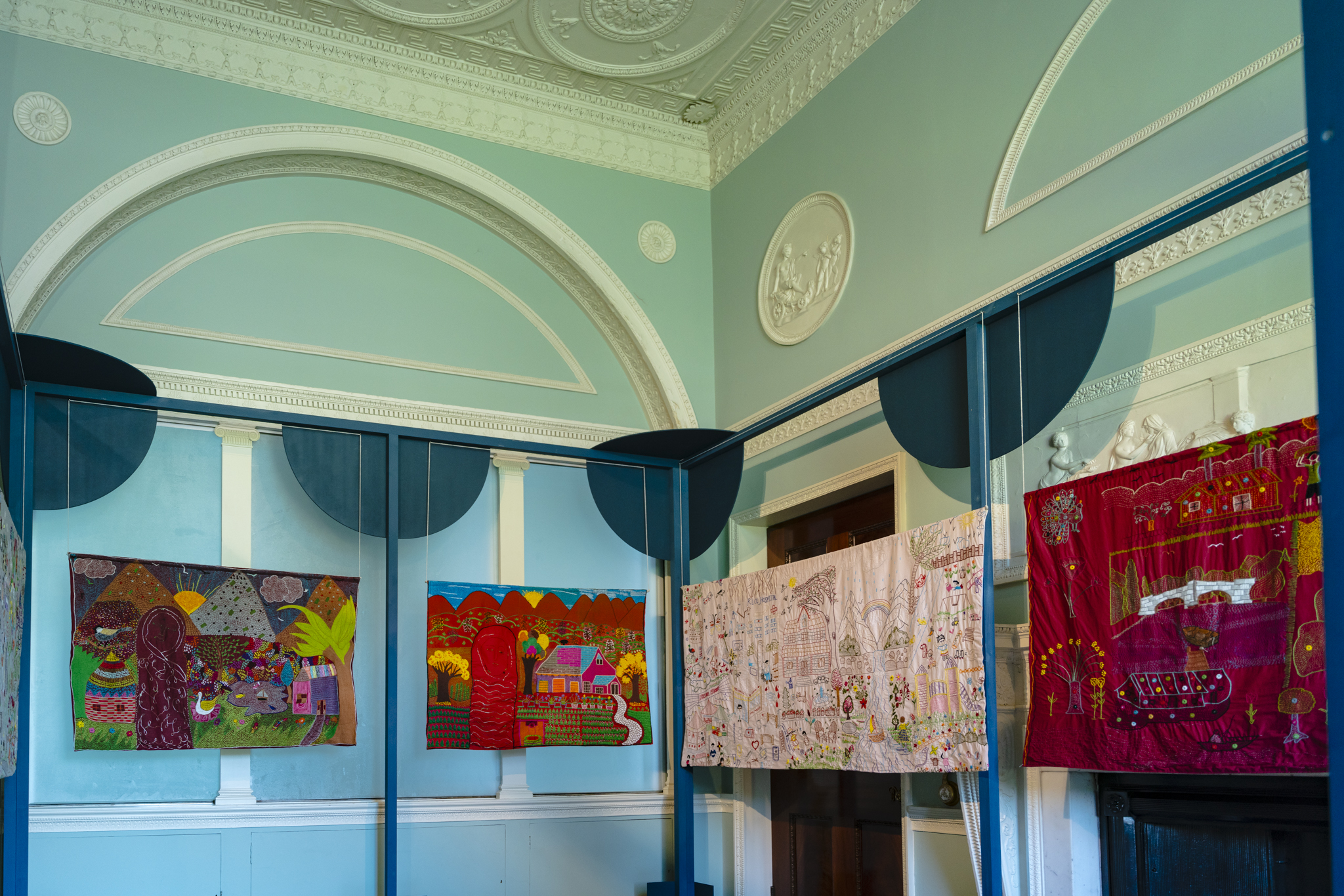 A series of embroidered panels created collaboratively by artisans from the Ra’ana Liaquat Craftsmen’s Colony (RLCC), women from the South Asian diaspora in Burnley and Bristol, and the Shantona Women’s Group in Leeds, hang from a dark blue frame. The colourful panels depict shared experiences of family and explore the meaning of home across different places and cultures.