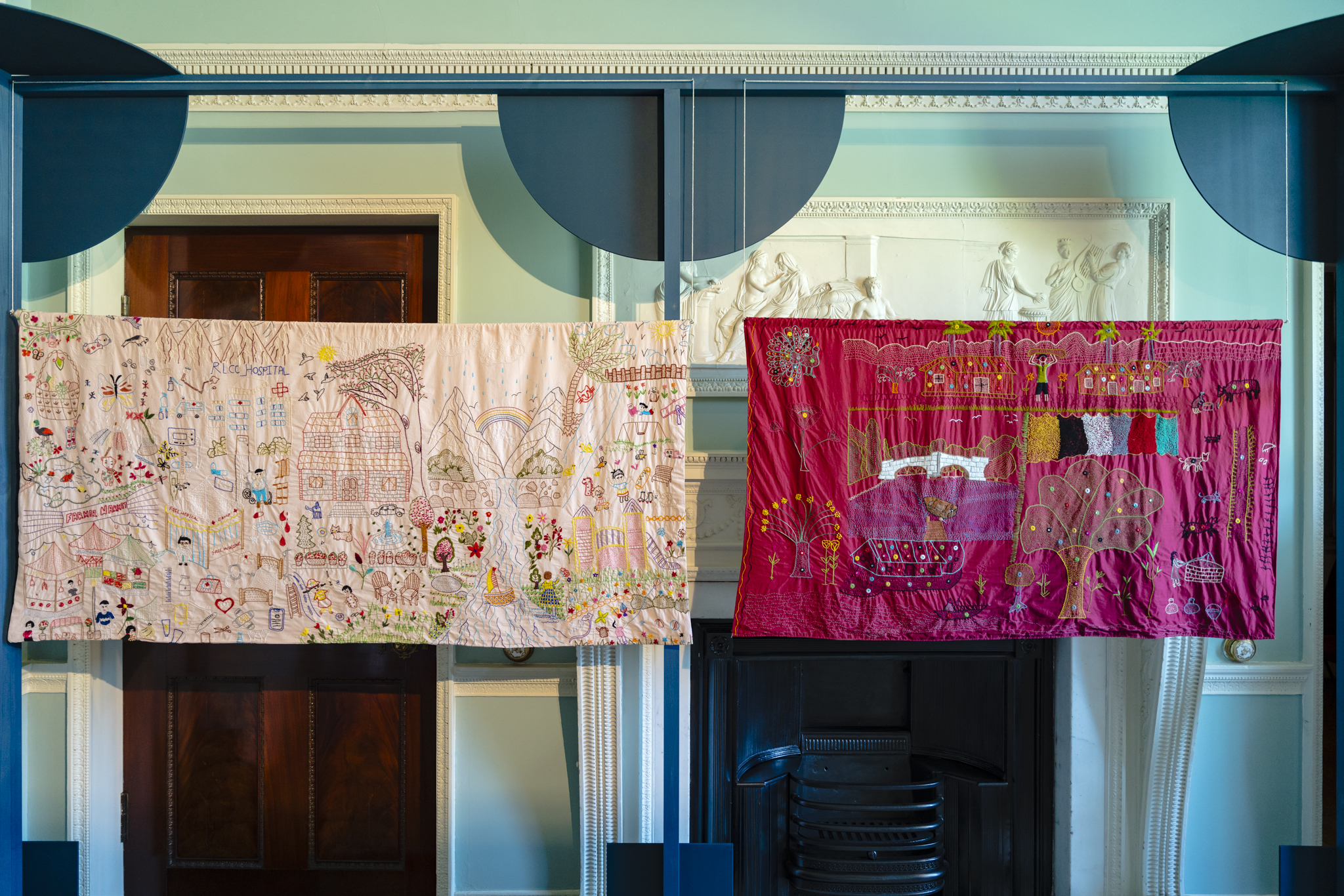 A series of embroidered panels created collaboratively by artisans from the Ra’ana Liaquat Craftsmen’s Colony (RLCC), women from the South Asian diaspora in Burnley and Bristol, and the Shantona Women’s Group in Leeds, hang from a dark blue frame. The colourful panels depict shared experiences of family and explore the meaning of home across different places and cultures.