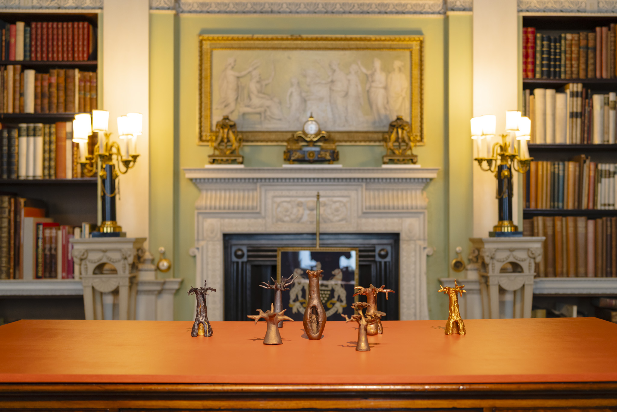 Seven miniature hand cast bronze Baobab trees stand proudly on a desk in the Old Library at Harewood. The desk is in the foreground, with a marble fireplace and book cases filled with vintage and rare books softly out of focus.