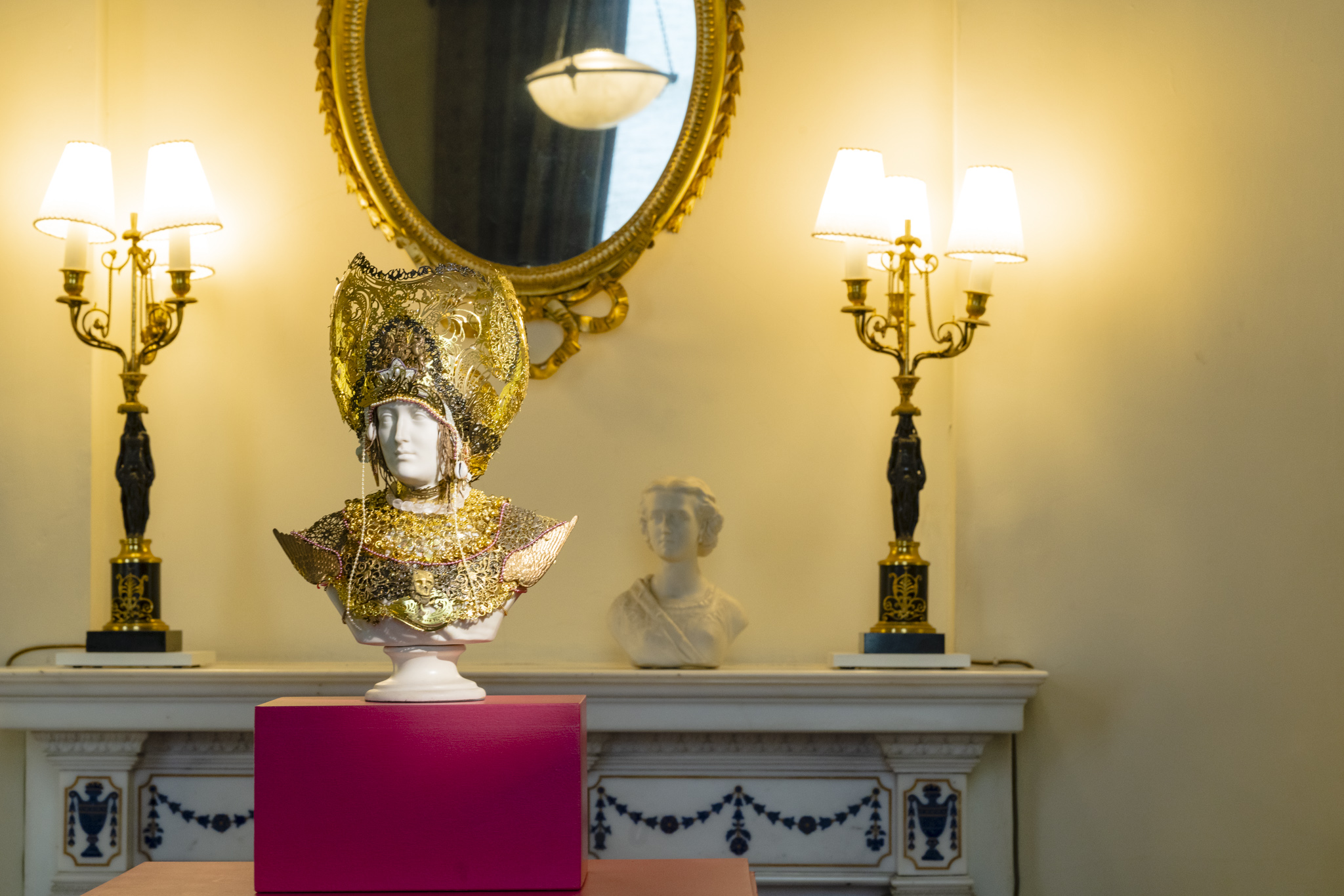 A small marble bust is adorned with a golden headdress stands on a bright pink display box. In the background, another marble bust sits on top of a marble fireplace, below a gilded Chippendale mirror.