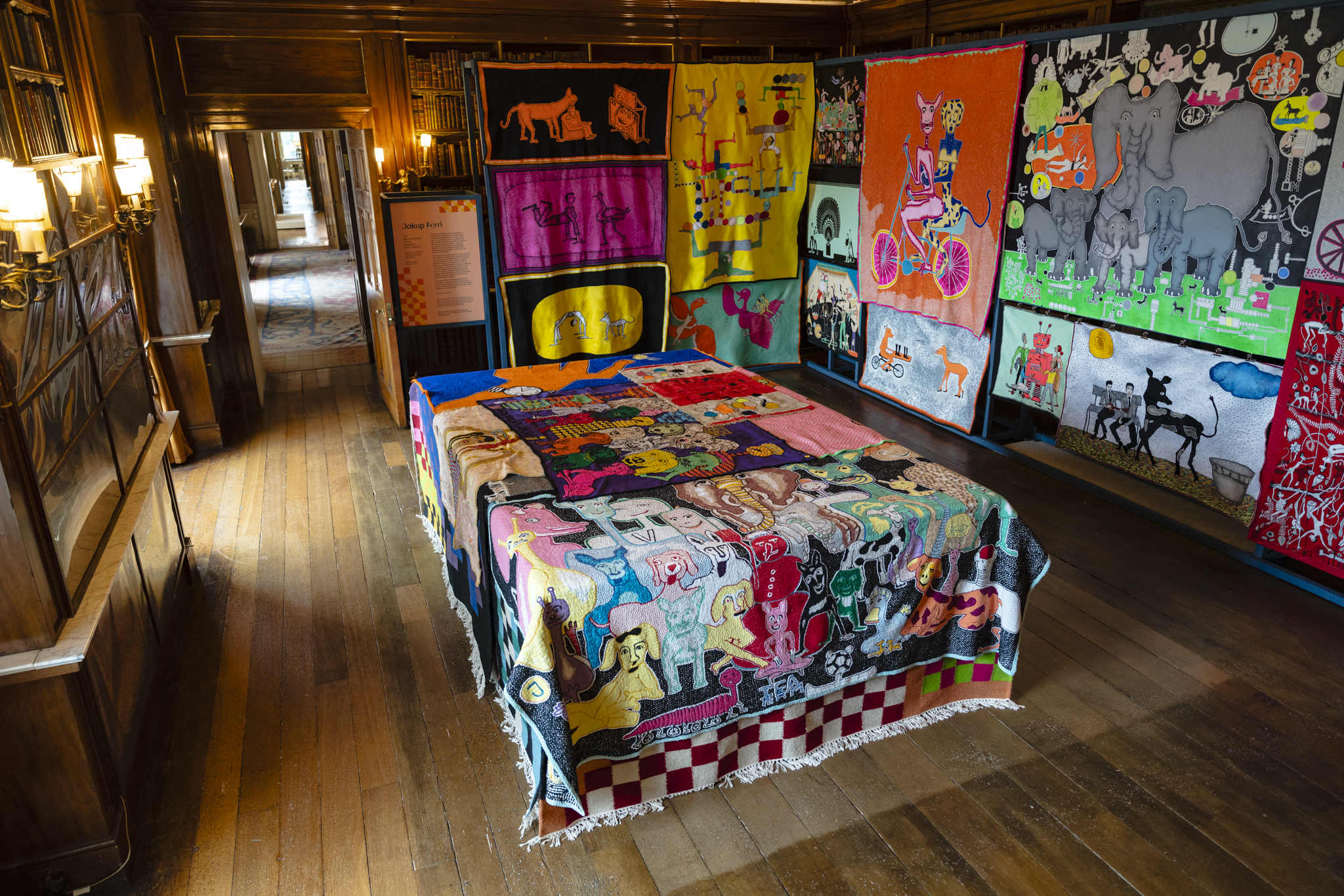 Brightly coloured hand made rugs cover the bookcases of the Spanish Library. Some rugs feature exaggerated cartoon style animals including elephants, dogs, cats and birds. In the centre of the room, a large box covered with even more hand sewn rugs.