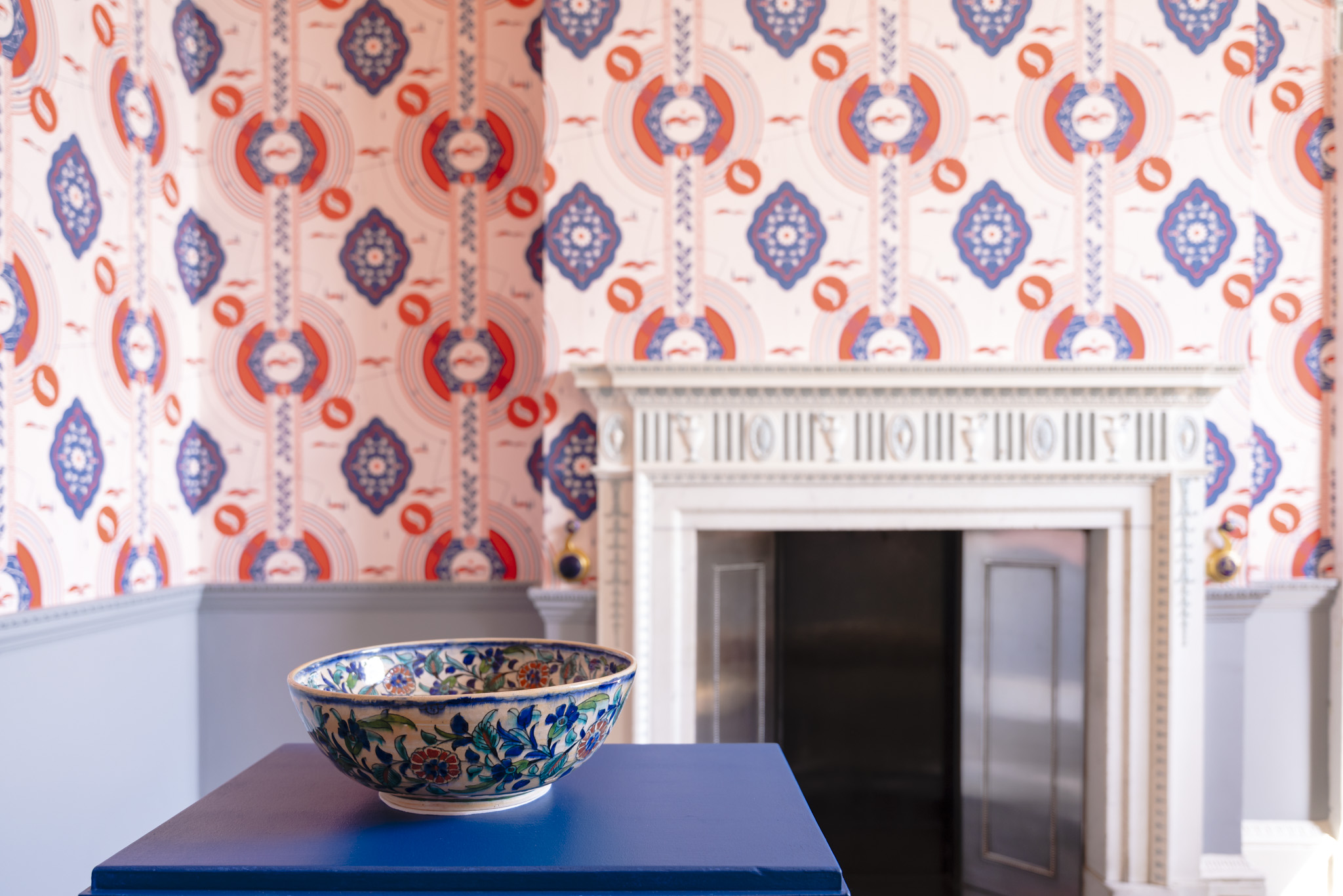 A blue floral Japanese china bowl sits upon a blue box in a small wallpapered room. The wallpaper has a repeated pattern of blue and red motifs inspired by the stories of those who worked and lived at Harewood House.