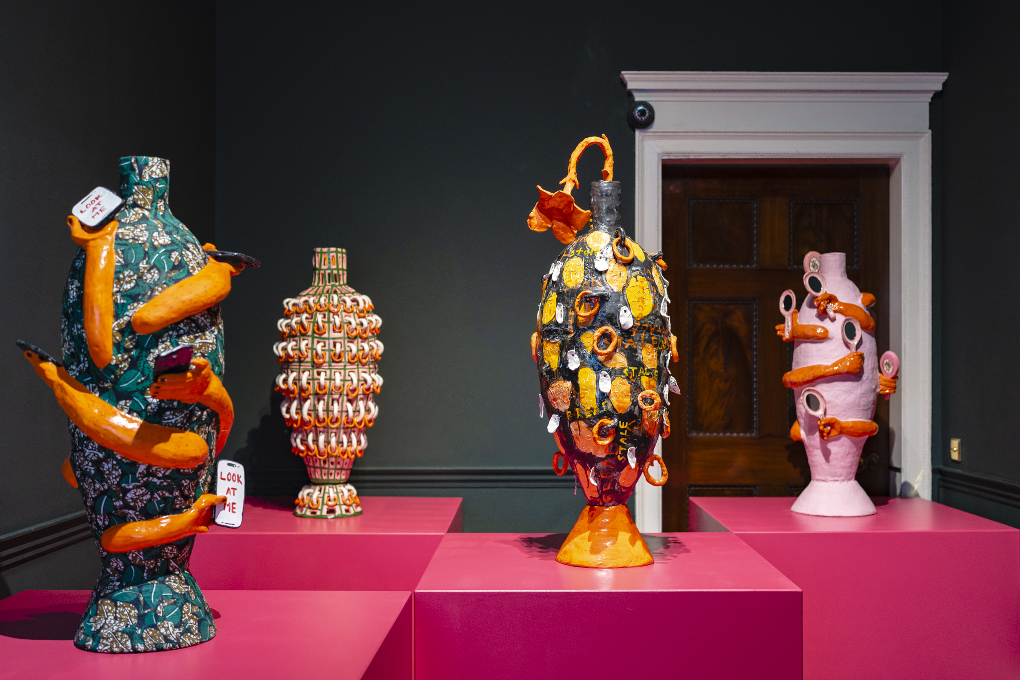 Four brightly coloured giant vases stand on top of rough hot pink boxes in a small dark room.