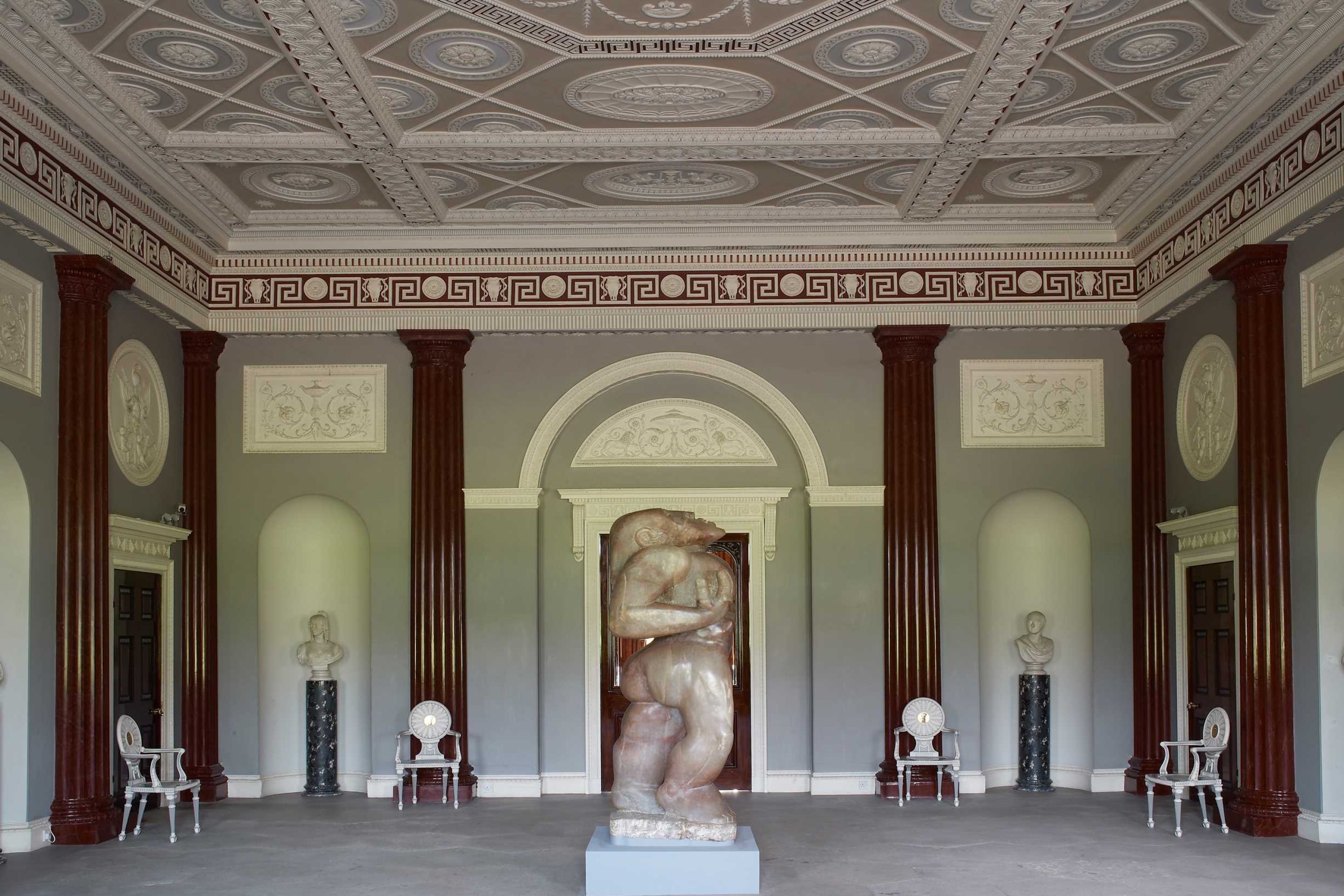 A grand room with faux-marble columns lining the walls and an intricate plasterwork ceiling painted in pastel colours. A large human alabaster sculpture stands in the centre of the room.