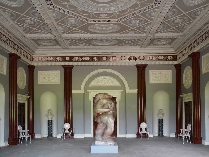 A grand room with faux-marble columns lining the walls and an intricate plasterwork ceiling painted in pastel colours. A large human alabaster sculpture stands in the centre of the room.