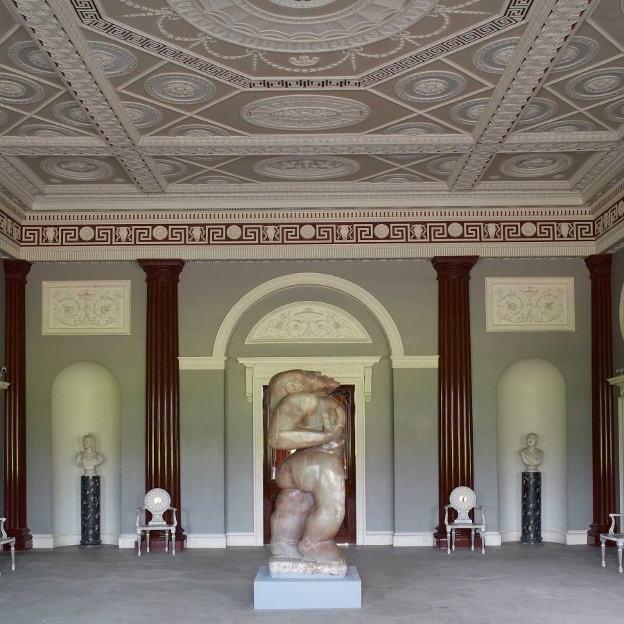 A grand room with faux-marble columns lining the walls and an intricate plasterwork ceiling painted in pastel colours. A large human alabaster sculpture stands in the centre of the room.