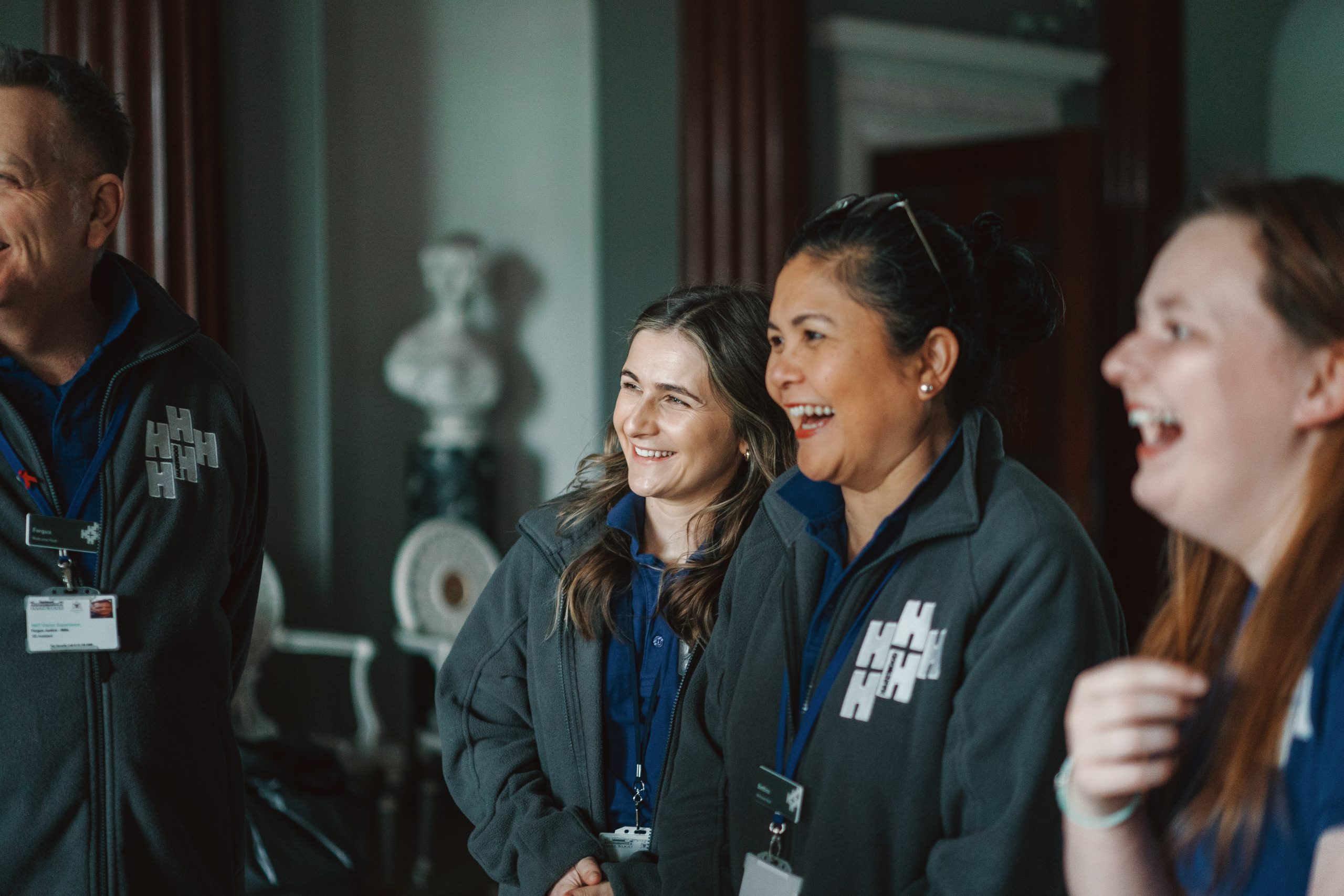 A group of happy Harewood staff standing in the Entrance Hall, wearing polo shirts and dark grey fleece jackets embroidered with the Harewood Logo.