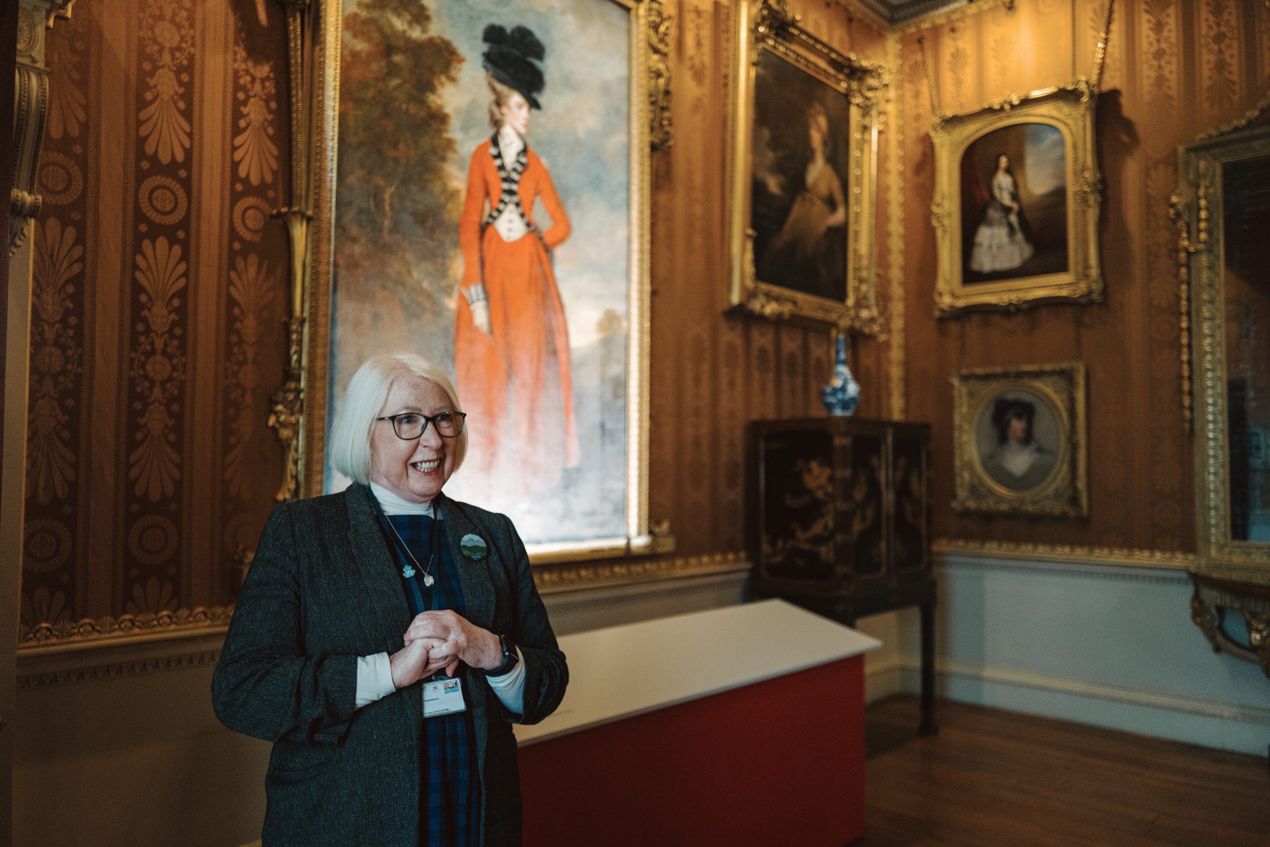 A friendly volunteer wearing a blue lanyard stands in the Cinnamon Drawing Room beneath a Reynolds painting of Lady Worsley ready to answer any questions visitors may have.