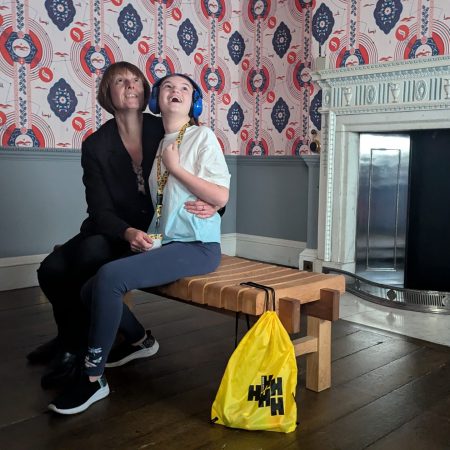 Adult and child sit on a wooden bench in Harewood Watercolour Room looking up at the intricate design of the red and blue hand block printed wallpaper. The child wears a sunflower lanyard and ear defenders. Harewood yellow sensory bag is at the end of the bench.