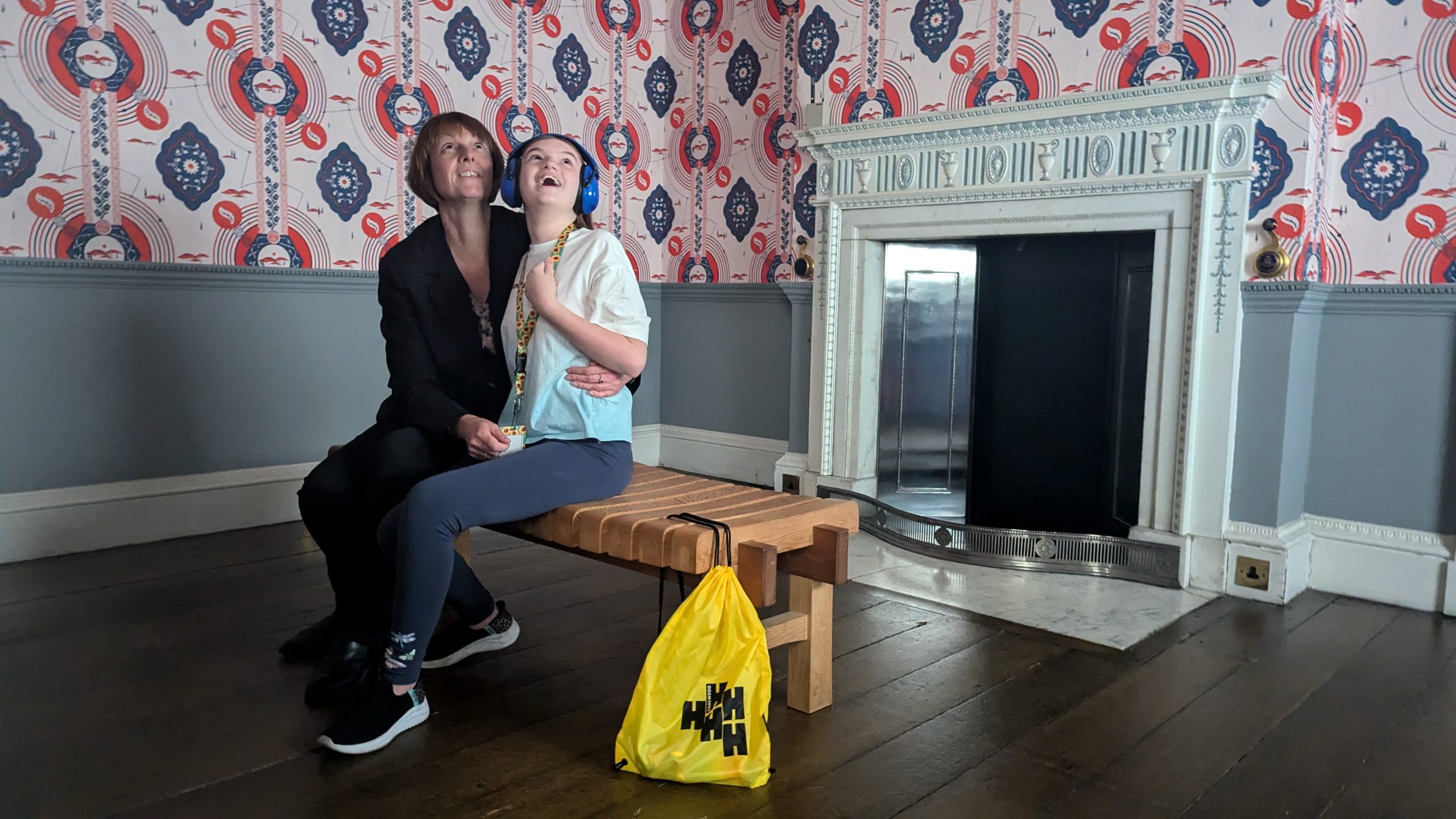 Adult and child sit on a wooden bench in Harewood Watercolour Room looking up at the intricate design of the red and blue hand block printed wallpaper. The child wears a sunflower lanyard and ear defenders. Harewood yellow sensory bag is at the end of the bench.