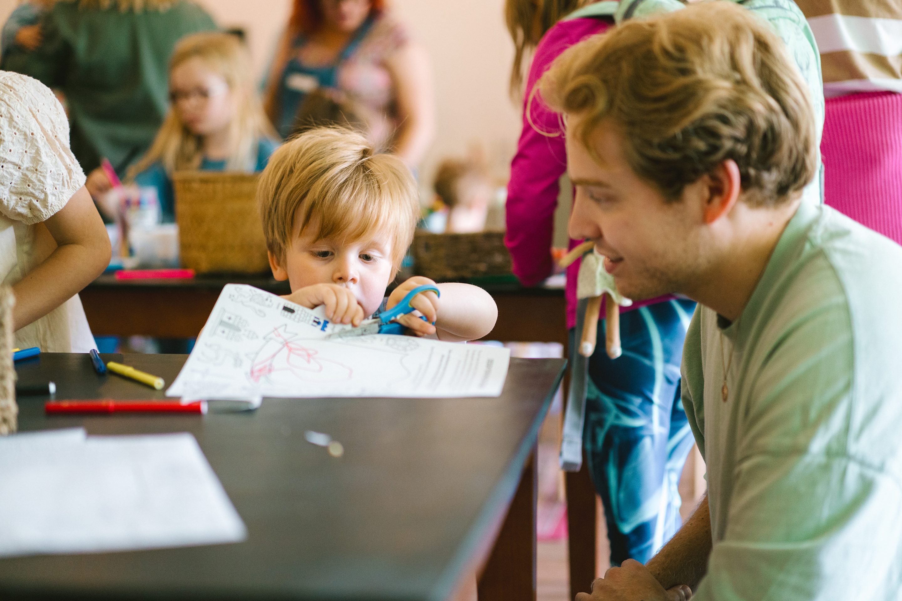 A young man oversees a young child using safety scissors to cut out a craft in the Servants' Hall.