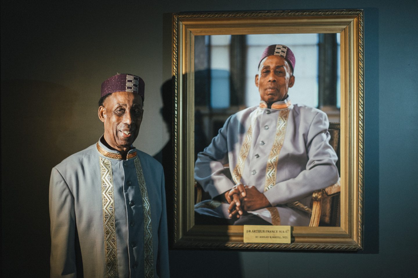 Dr Arthur France MBE stands next to a framed portrait of himself. Both wear a traditional embroidered light grey suit and a patterned purple hat, exuding pride and dignity.