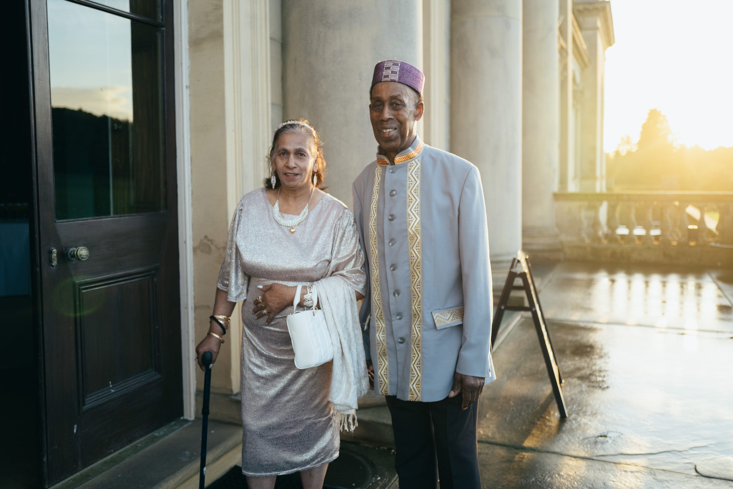 An elderly couple stands smiling outside a grand building at sunset. The woman wears a shimmering silver dress and uses a walking stick; the man wears a traditional embroidered outfit with a purple hat.