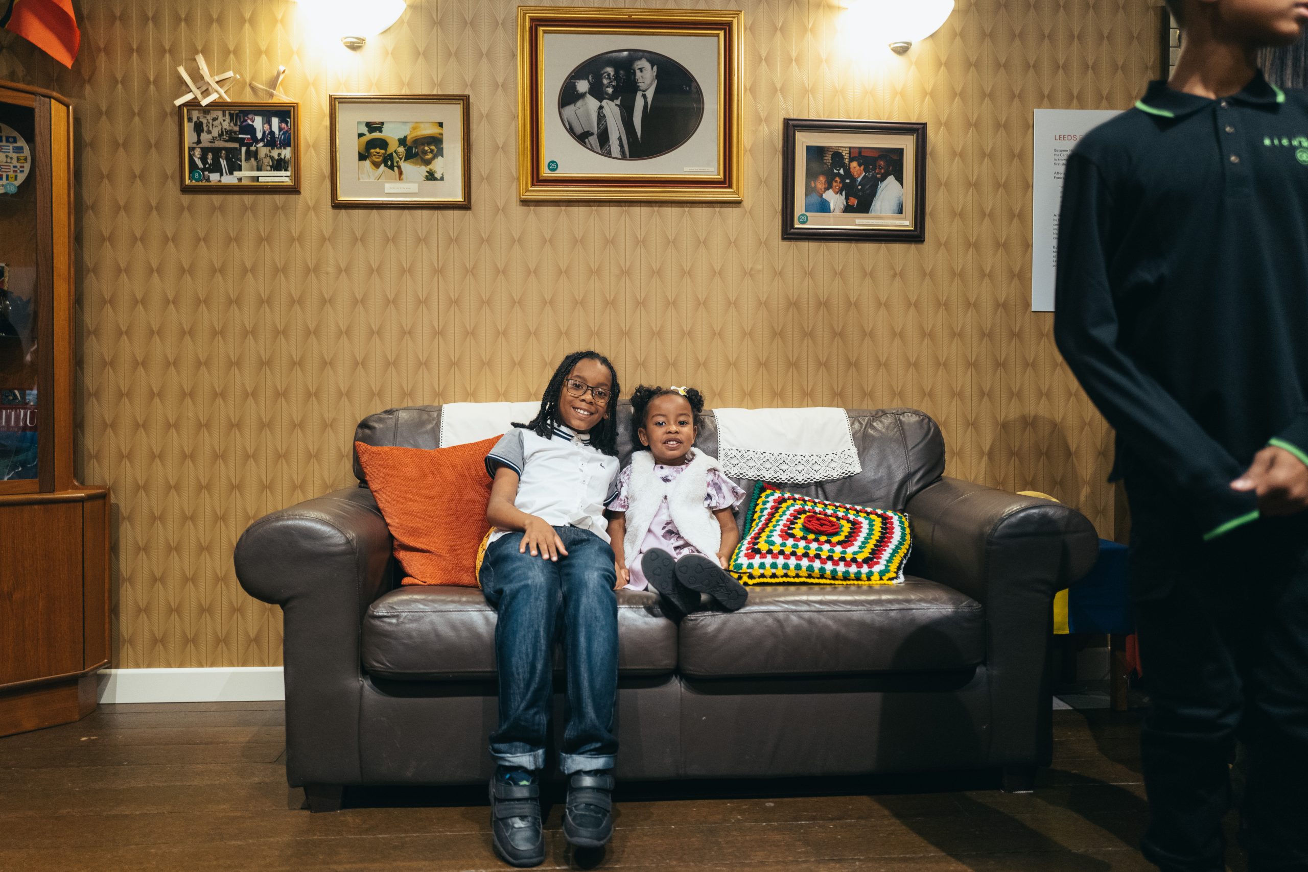 A boy, and a young girl sit on a grey leather sofa in a room recreated to look like Arthur France's living room.