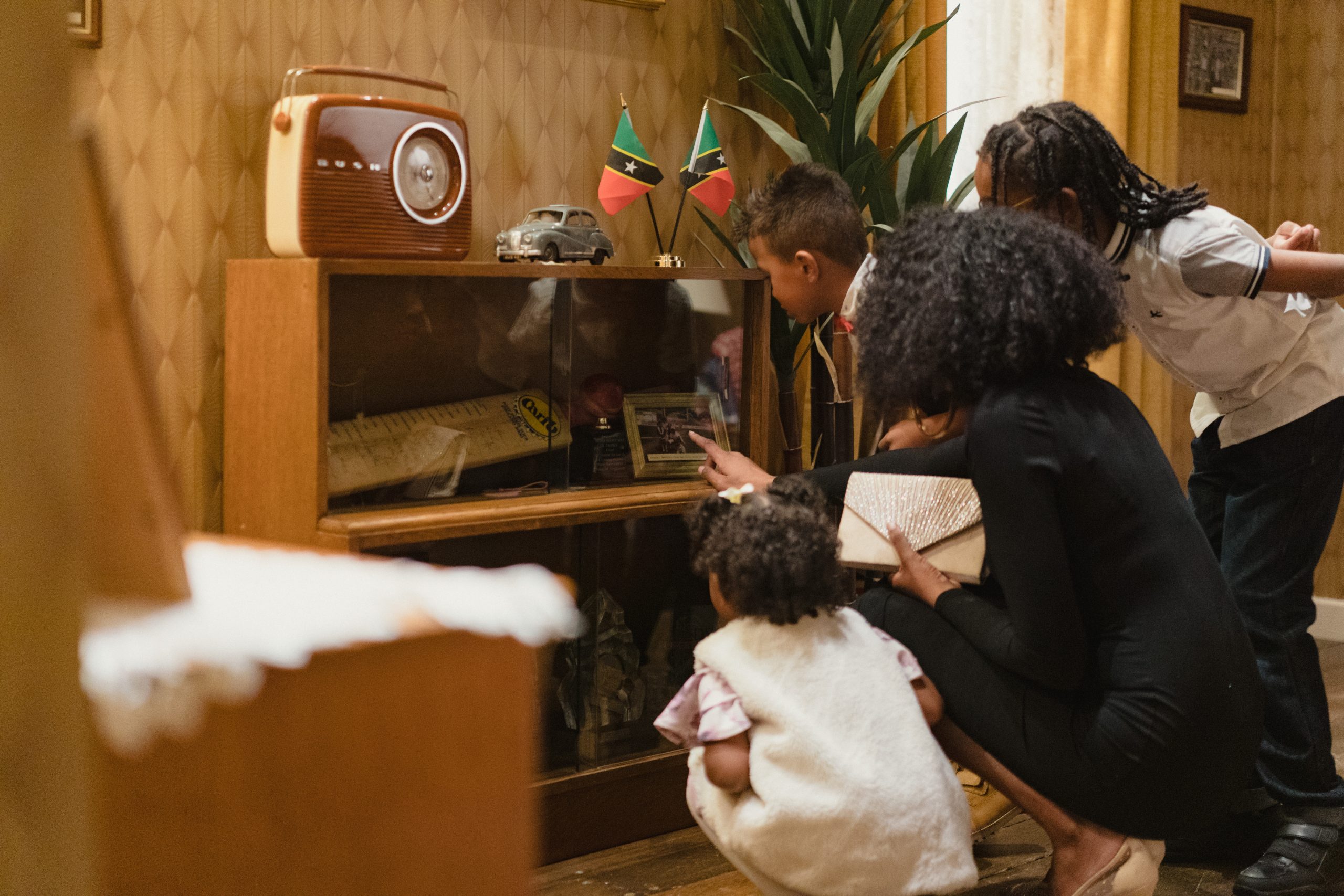 A family gathers around a vintage cabinet, viewing memorabilia. A woman holds a small gold clutch bag, while two children lean in. The setting feels warm and nostalgic.