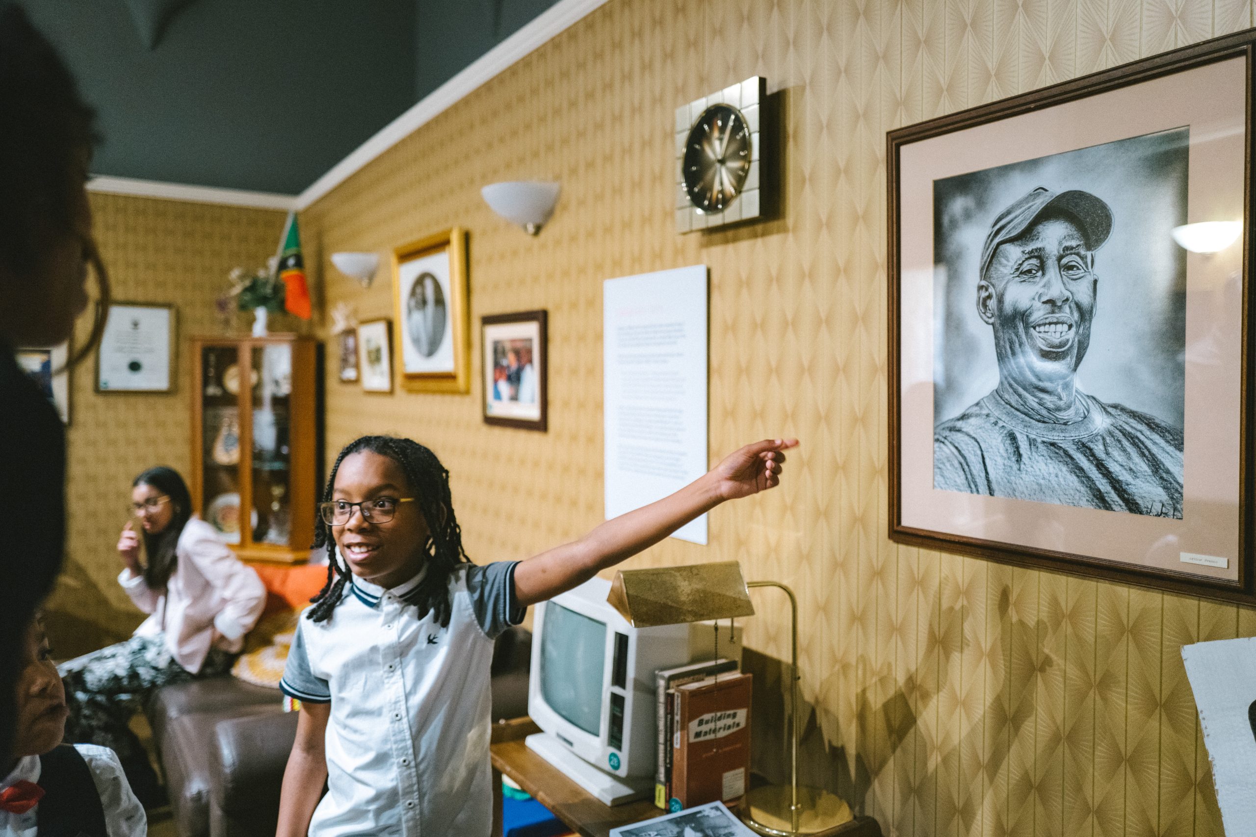 A child with shoulder length braids points with intrigue at a hand drawn picture, in a frame hung on a wall, of a man wearing a t shirt and baseball hat.