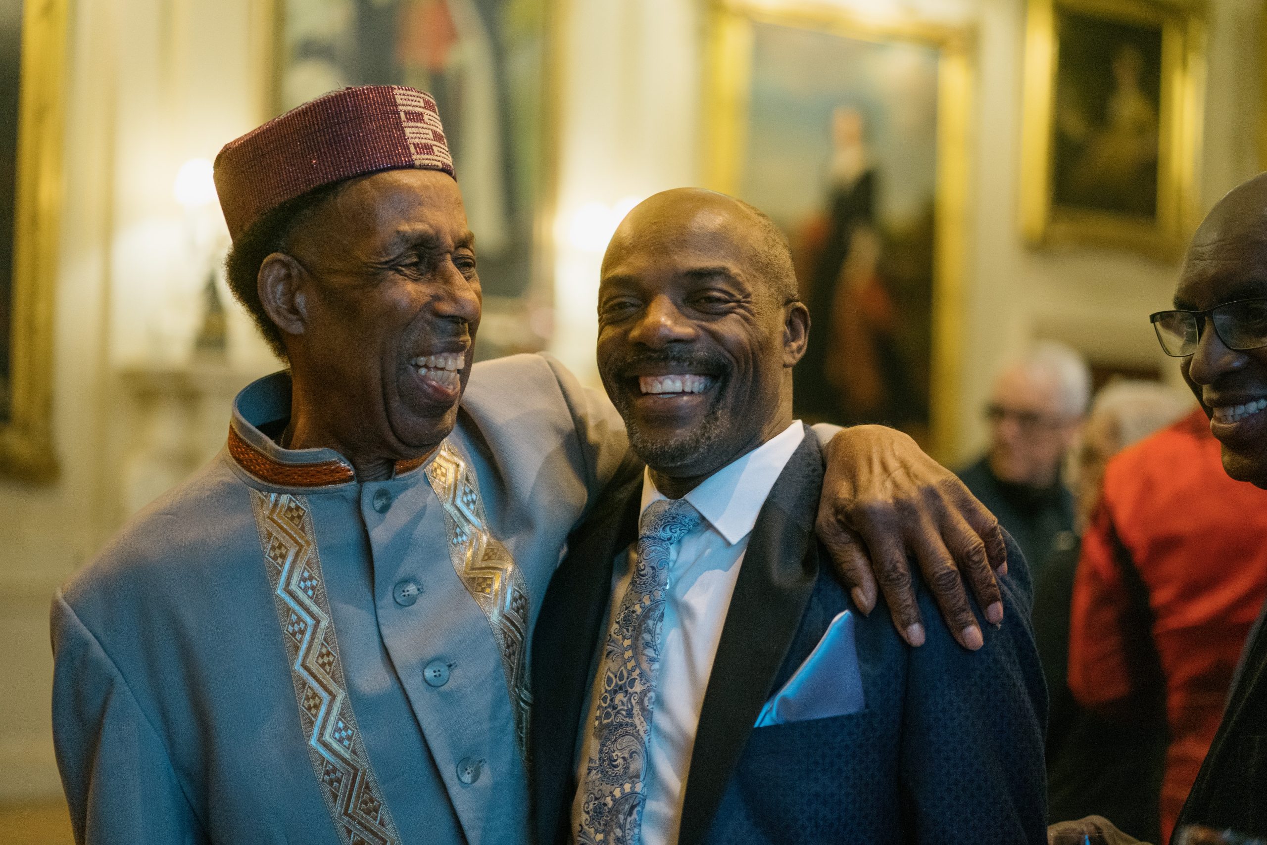 Two very happy men, laugh together with their arms around each others shoulders. One wears traditional attire with a patterned purple hat, the other a blue suit with white shirt and light blue paisley tie.