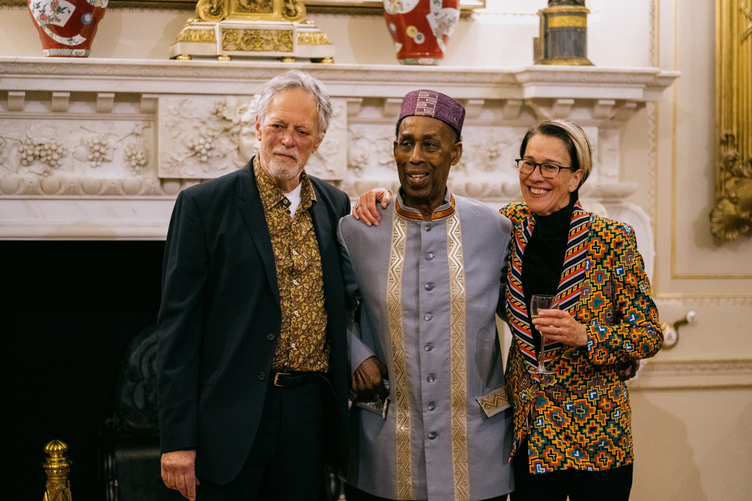 Three people stand in front of a large marble fireplace. On the left, David Lascelles, Earl of Harewood. David is a tall man with grey hair. He wears a black blazer over a ditsy leaf pattered shirt with white t shirt underneath. In the middle, Dr Arthur France smiles warmly, wearing a detailed gray and gold traditional outfit with a purple hat. On the right, Dianne Lascelles, Countess of Harewood wears glasses and a vibrant, geometric-patterned jacket.