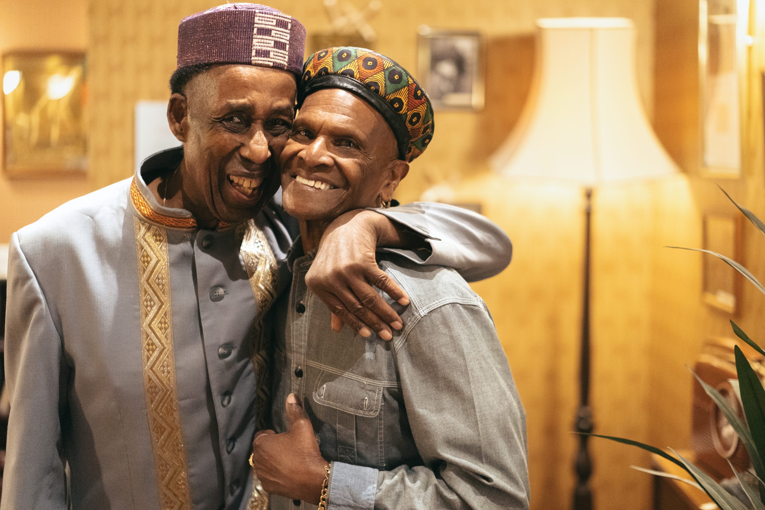 Two elderly men embracing warmly, wearing colourful hats and traditional clothing, smiling joyfully. The setting is warm and cosy with a lamp in the background.