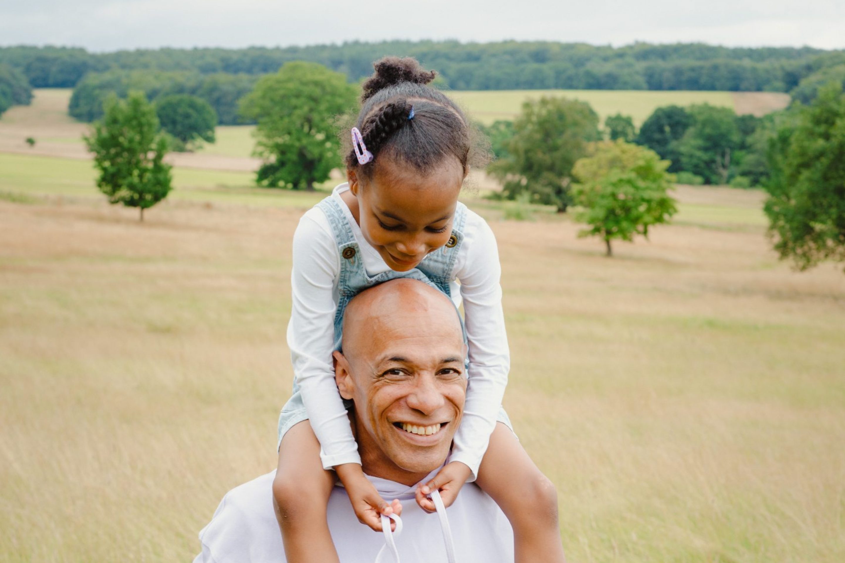 A smiling man in a light coloured hoodie carries a young girl on his shoulders through a grassy field. The girl, wearing pink adidas trainers and a denim dress, looks down playfully. The background features lush greenery and a cloudy sky.