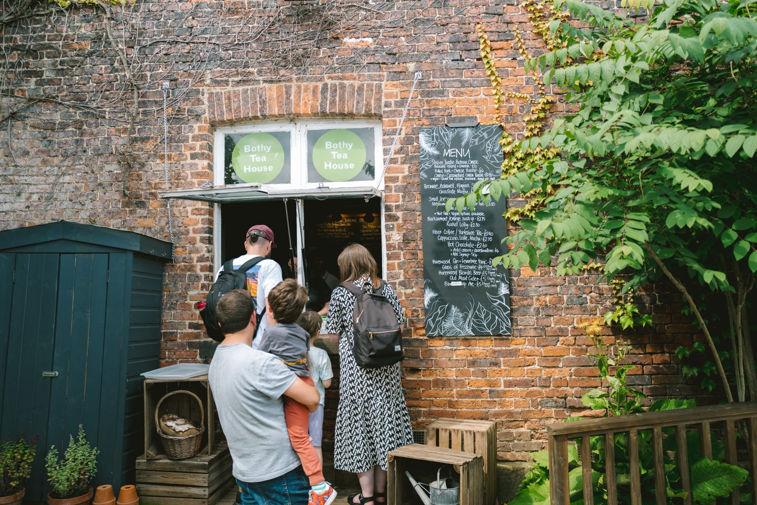 Two families queue outside an open window at the side of an old brick building. A blackboard lists menu items on one side with a small wooden displace cabinet on the other.