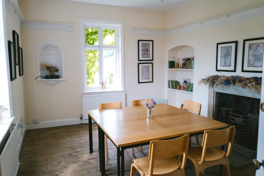 A square wooden table and four chairs stand in the middle of a small room. Dried flowers lay across a mantlepiece above a cast iron fireplace. Light trickles in through a window on the far wall.