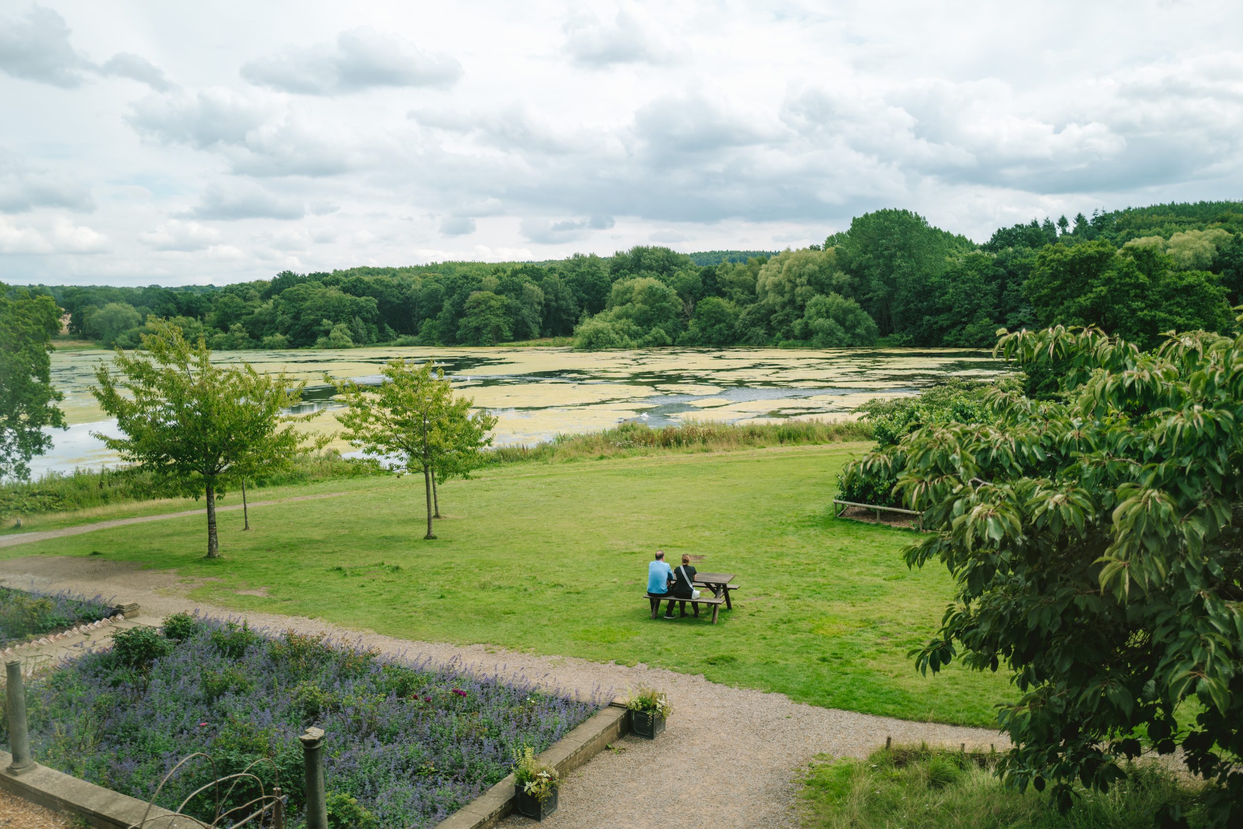 Vibrant green trees huddle around the edges of the Harewood Lake under a dramatic cloudy sky. Two people sit on the picnic bench outside the Bothy Teahouse, next to the Lavender and Rose Garden.