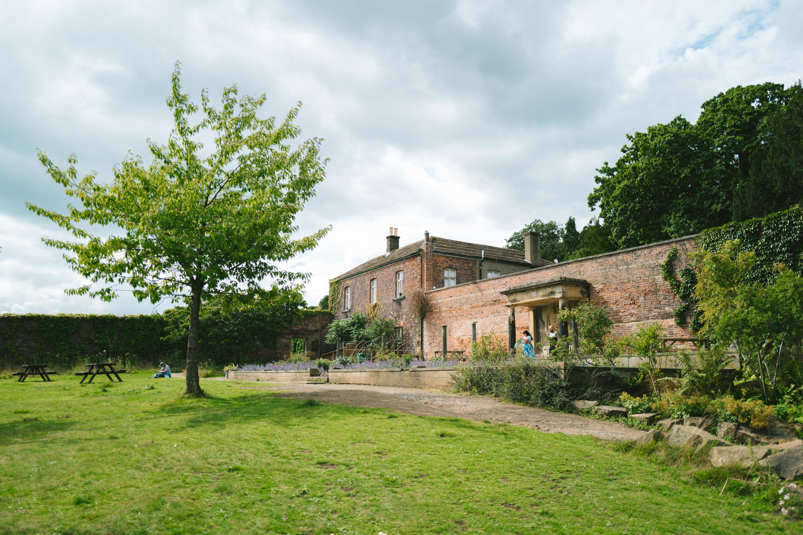 People stand amongst the Lavender and Rose Garden above the Bothy lawn. The the background, a person sits on the grass outside the Walled Garden waiting for a hot chocolate from The Bothy Teahouse.