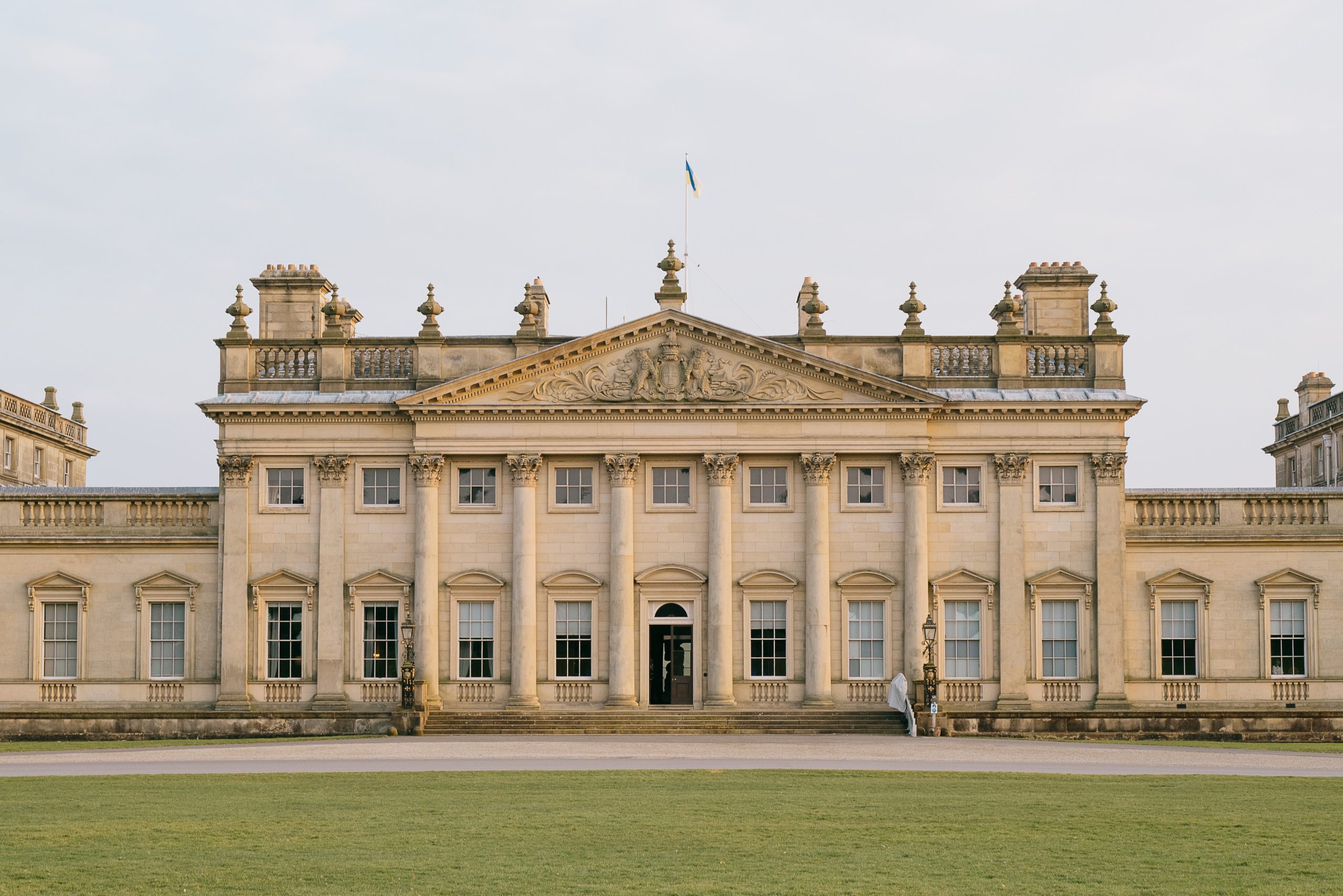 Front view of Harewood House showcasing the detailed architecture designed by York architect John Carr in the Palladian style. A grand, symmetrical façade with a central portico supported by six classical columns, large sash windows, and intricate stone detailing. Stone staircase leads up to the front door which is open, inviting visitors to enter in.